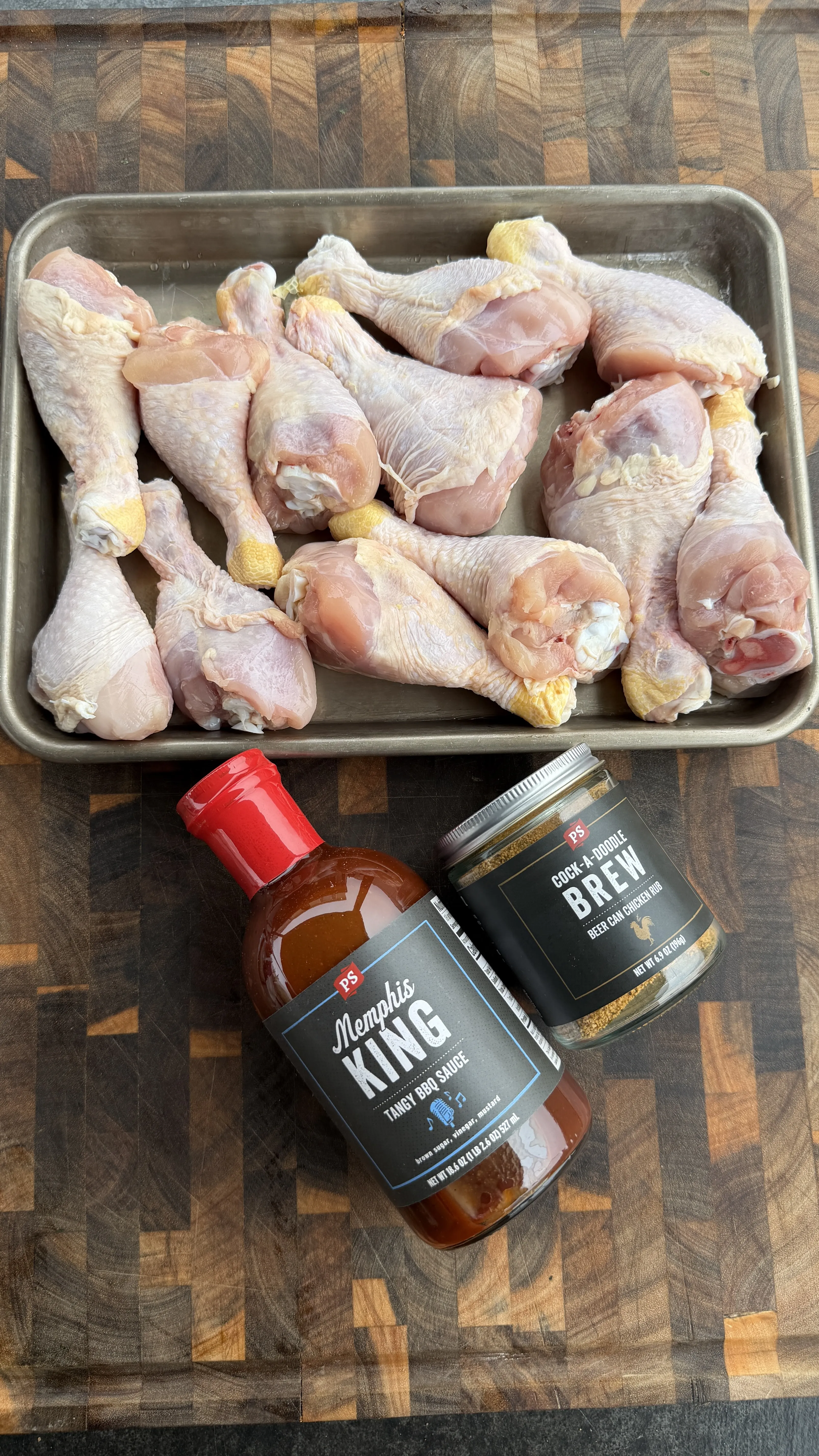 Raw chicken drumsticks in metal tray with barbecue seasonings on wood cutting board