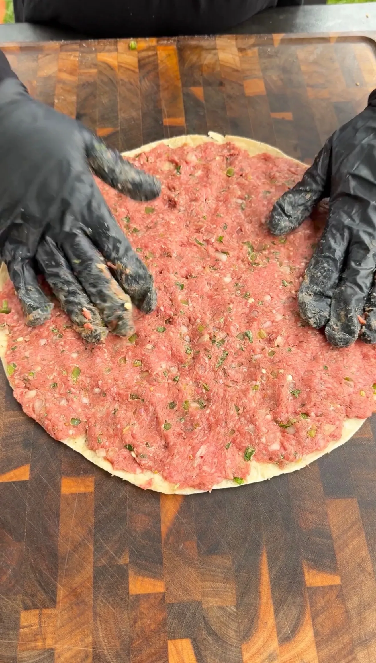 Hands in black gloves spreading seasoned raw ground beef mixture with herbs on flour tortilla for pinwheels