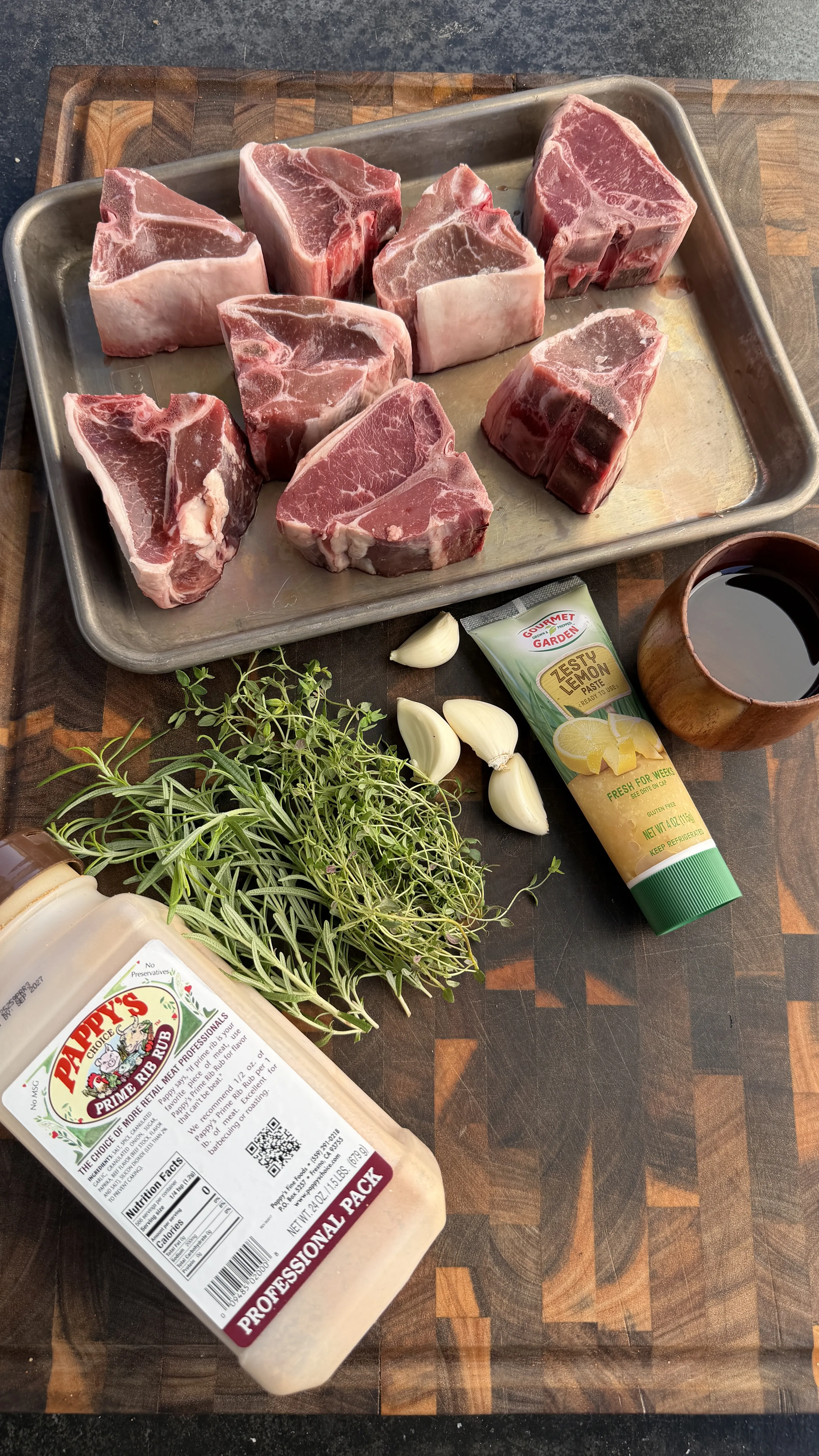 Raw lamb loin chops on metal tray with fresh herbs, garlic cloves, and seasonings on wooden cutting board