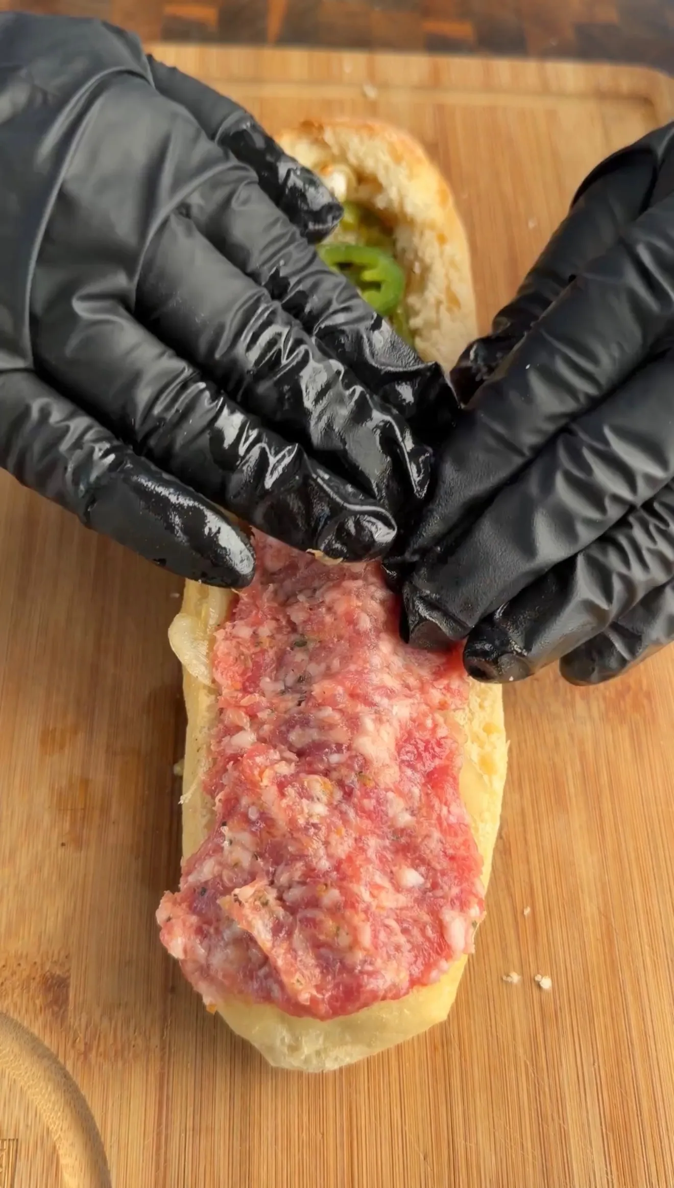 Hands in black gloves spreading raw sausage meat into hollowed bread boat on wooden cutting board