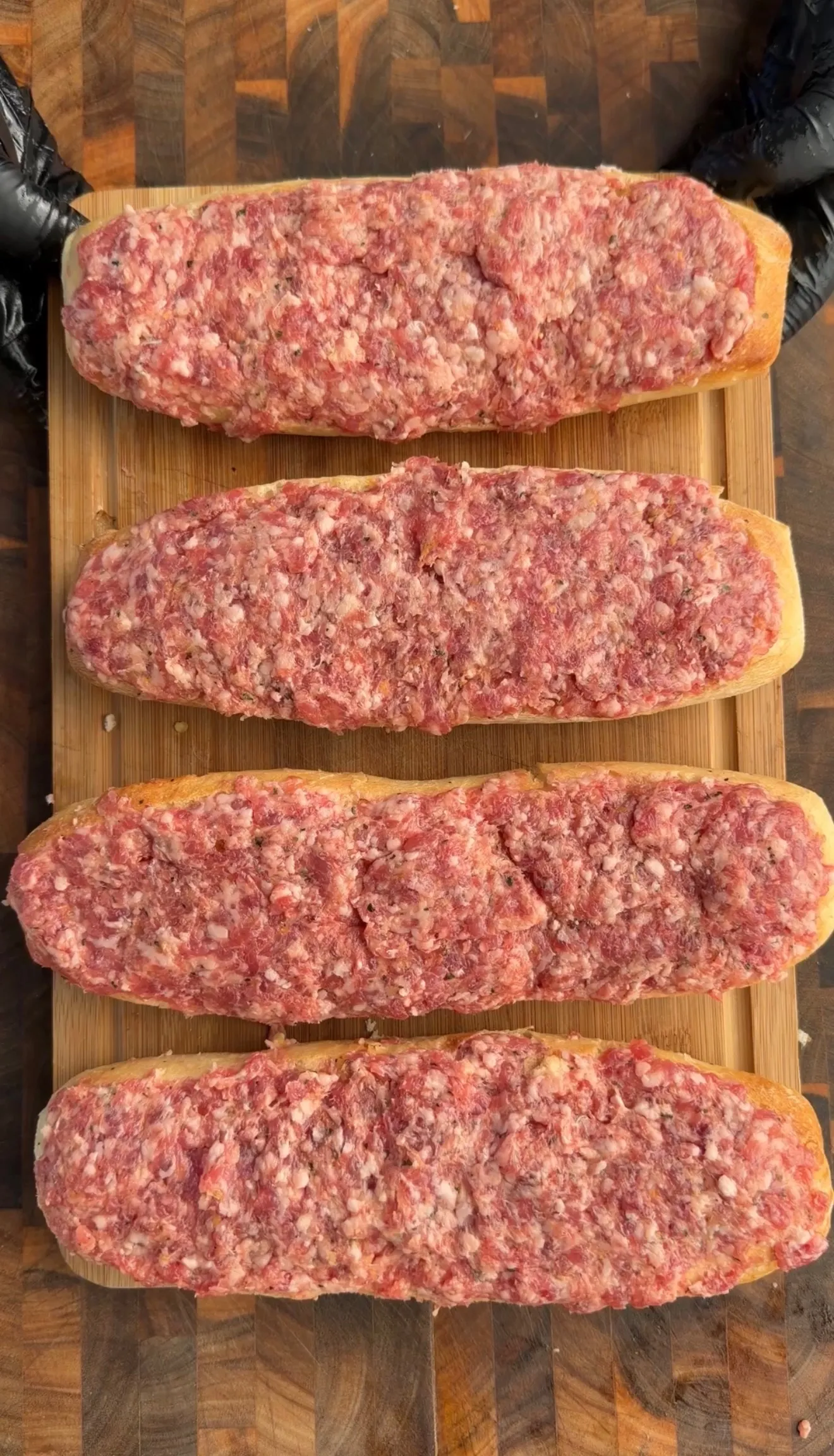 Four uncooked bread boats filled with raw sausage meat mixture arranged on a bamboo cutting board