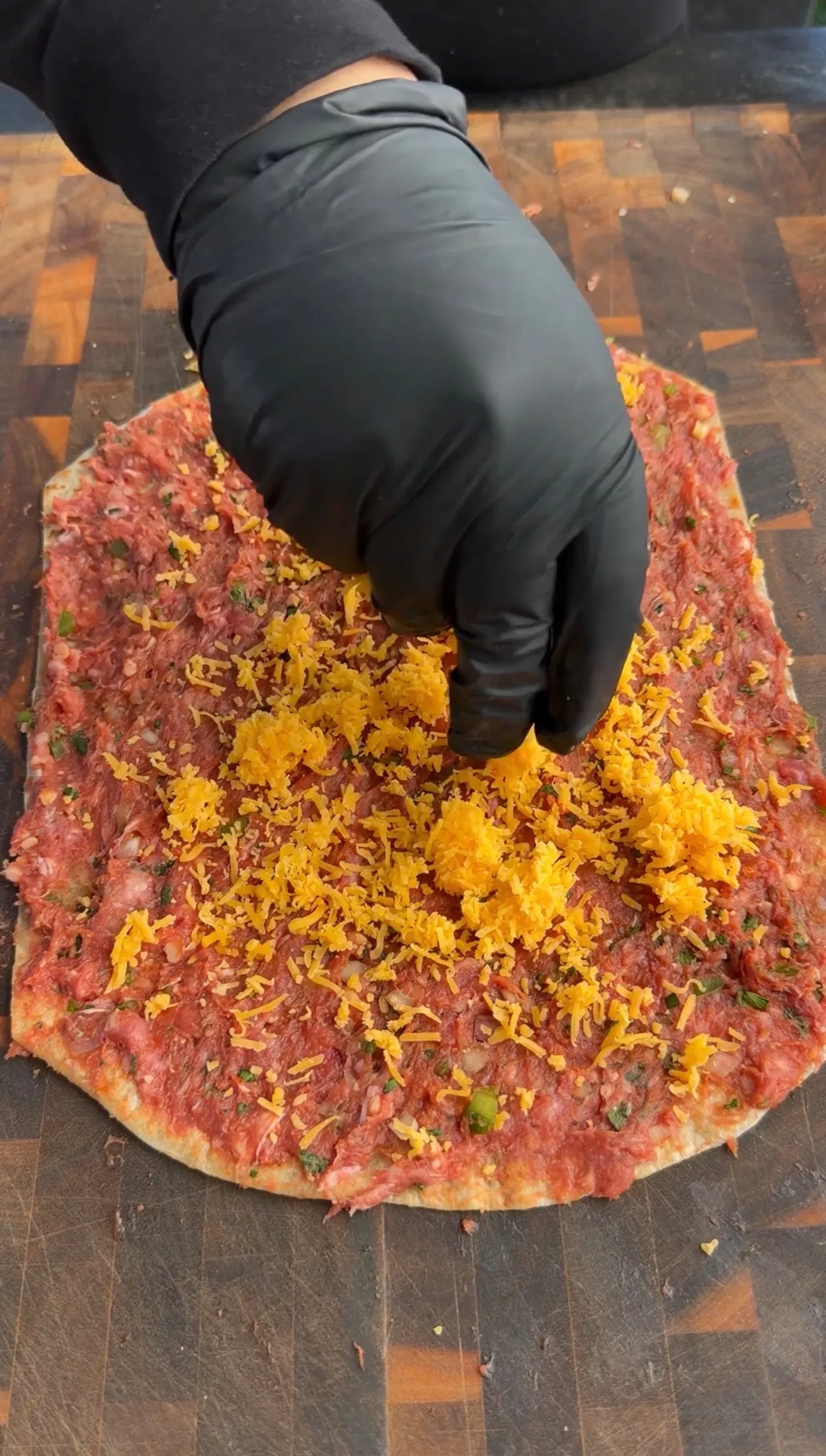 Raw seasoned ground lamb spread on tortilla with shredded cheese being added