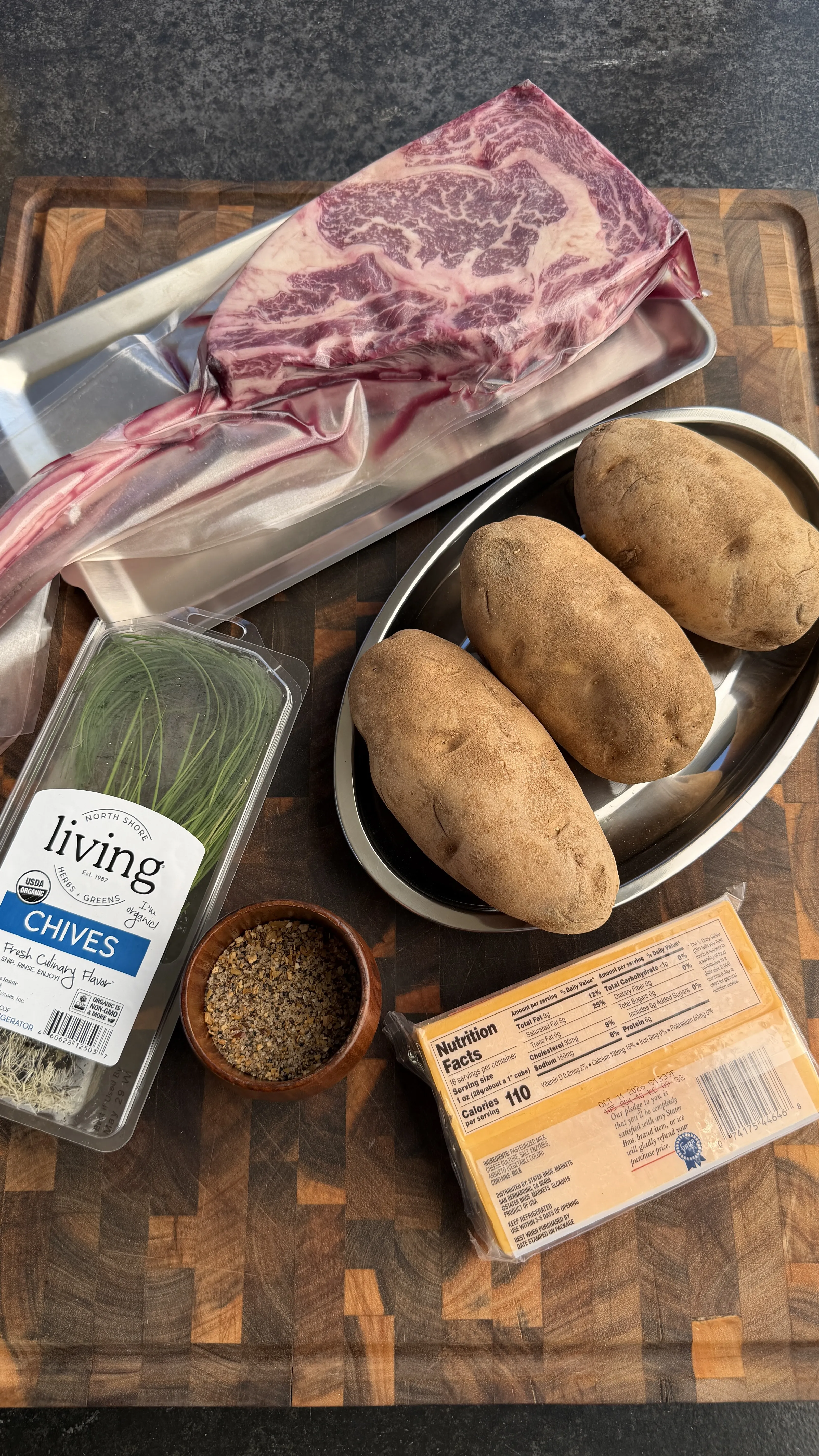 Raw tomahawk steak with potatoes, chives, cheese, and seasonings arranged on wooden cutting board