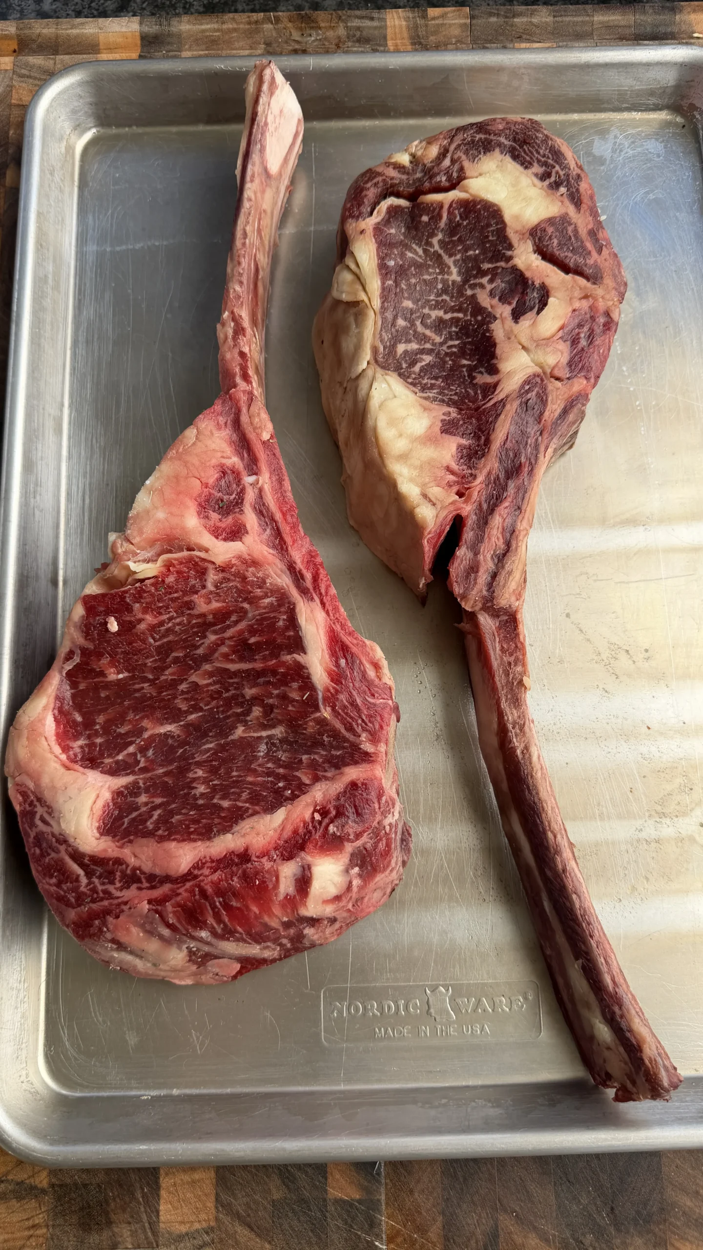 Two raw tomahawk steaks with long bones on a stainless steel baking tray, ready for salt block searing