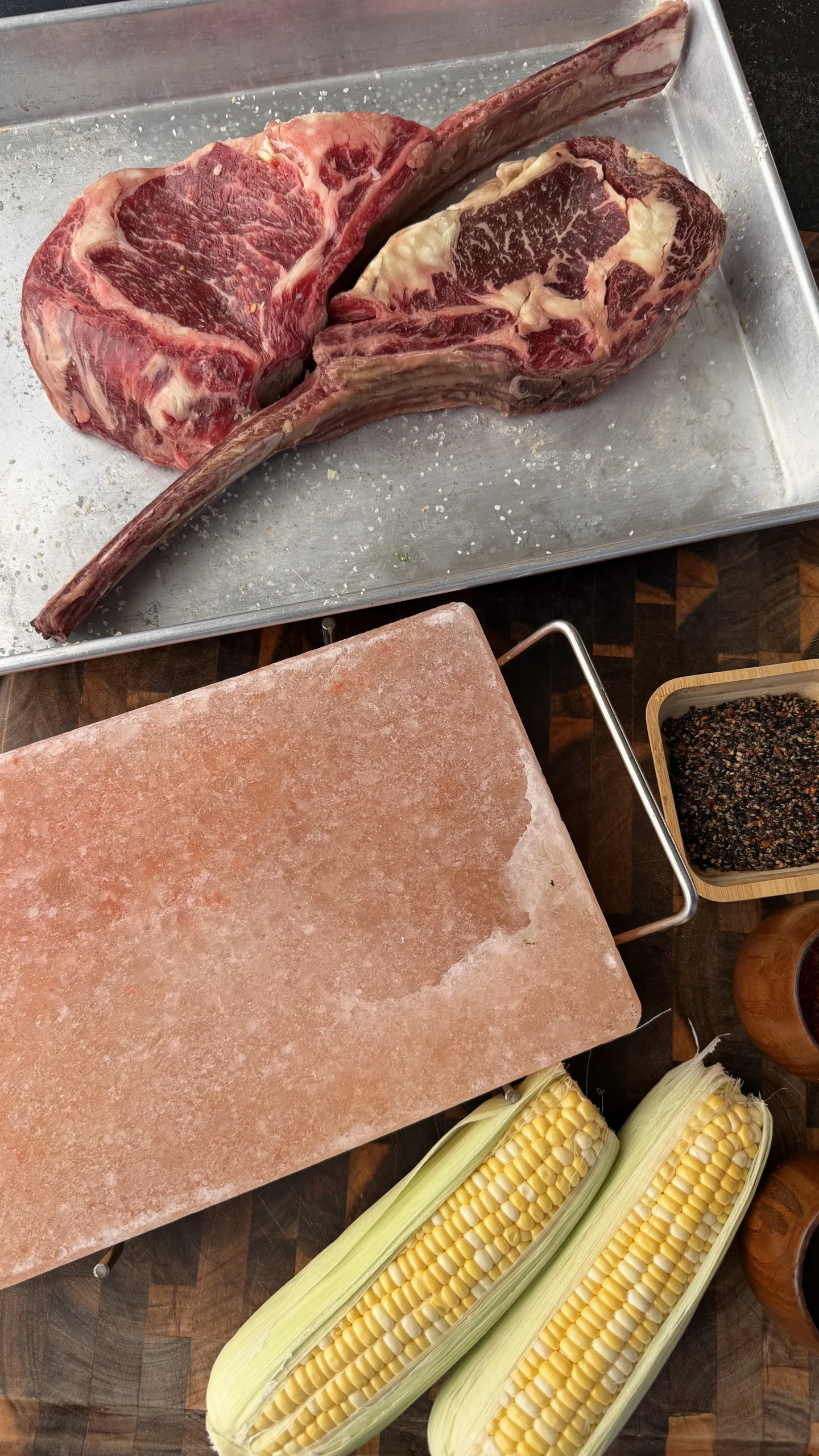 Two raw tomahawk steaks on metal tray with pink salt block, corn ears, and seasoning bowl for grilling prep