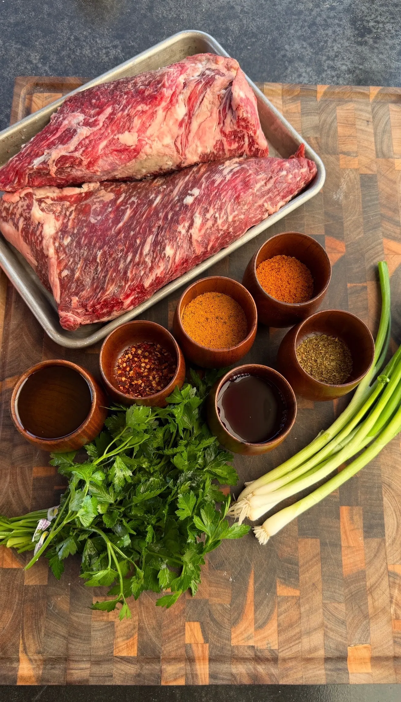Raw tri-tip roast on metal tray with spice bowls, fresh herbs, and green onions on wooden cutting board
