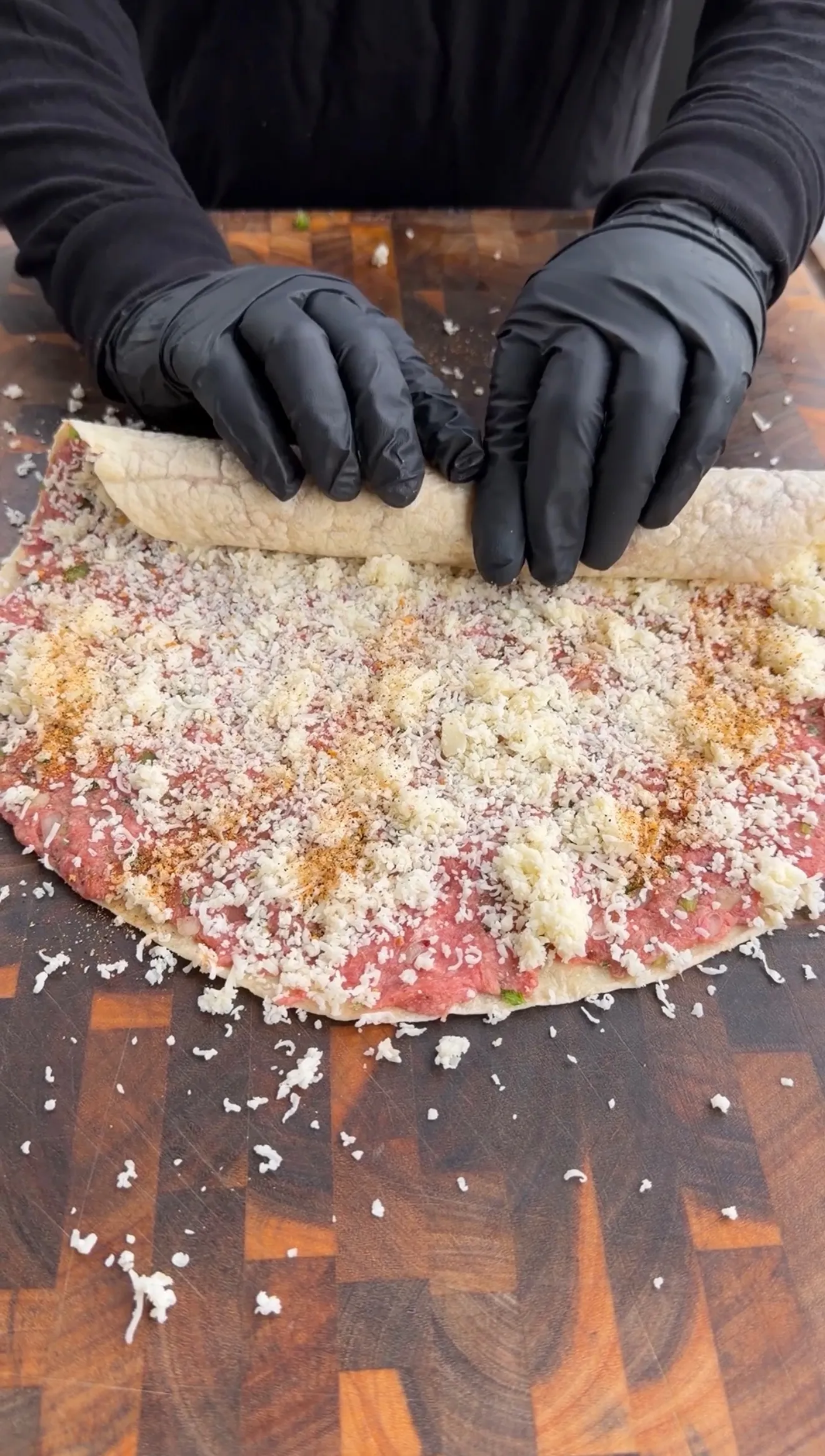 Hands in black gloves rolling cheesy beef tortilla pinwheels covered with grated cheese on wooden surface