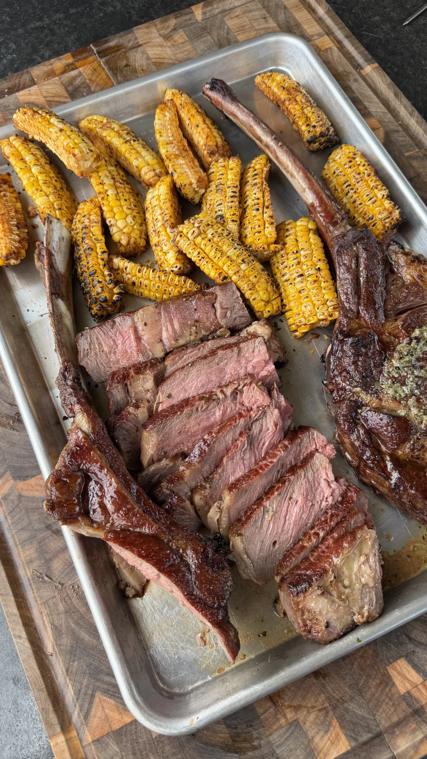 Sliced tomahawk steak and grilled mini corn ribs on a metal sheet pan on wooden cutting board