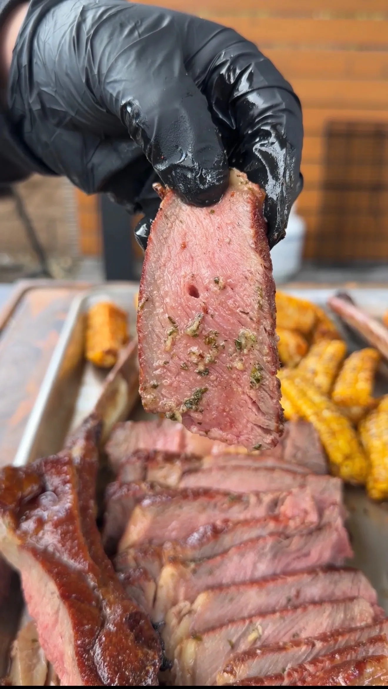 Hand holding slice of medium-rare salt block seared tomahawk steak with herbs, corn ribs visible on cutting board