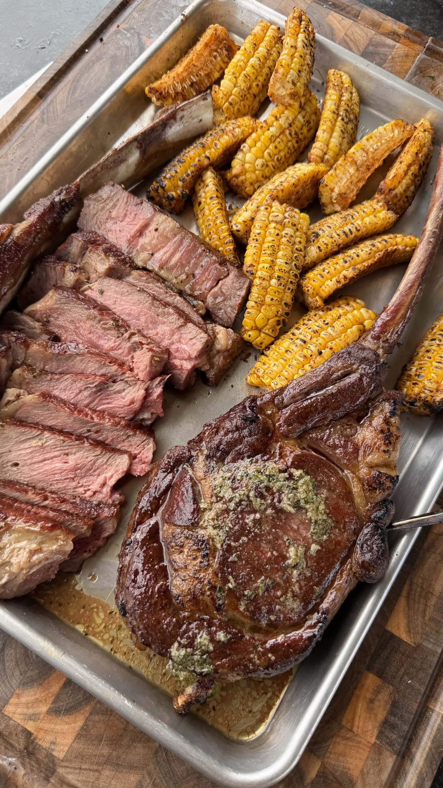 Sliced tomahawk steak with herb butter and grilled mini corn ribs on a metal serving tray