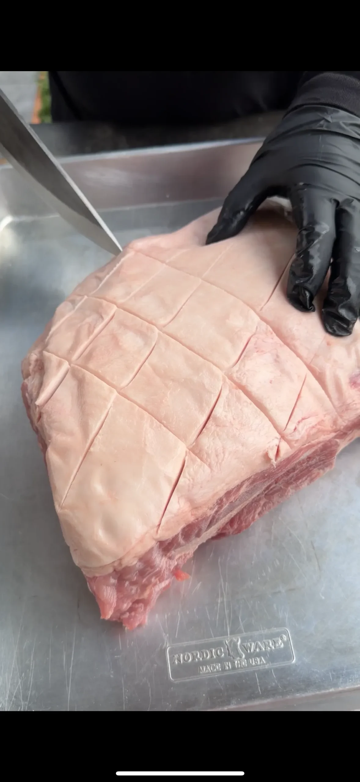 Raw pork shoulder with crosshatch scoring pattern on cutting board, being prepared by chef in black gloves