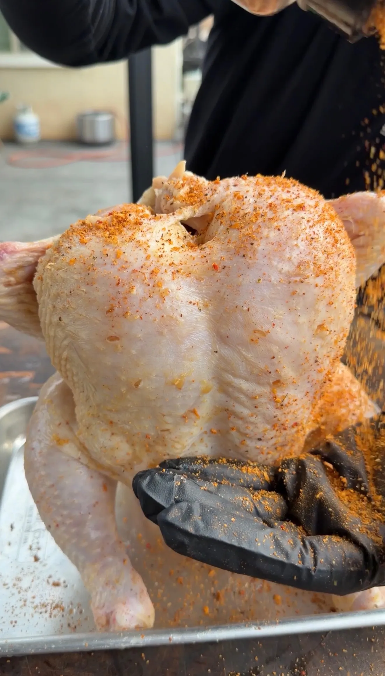 Raw whole chicken being seasoned with orange spice rub by chef wearing black gloves in kitchen