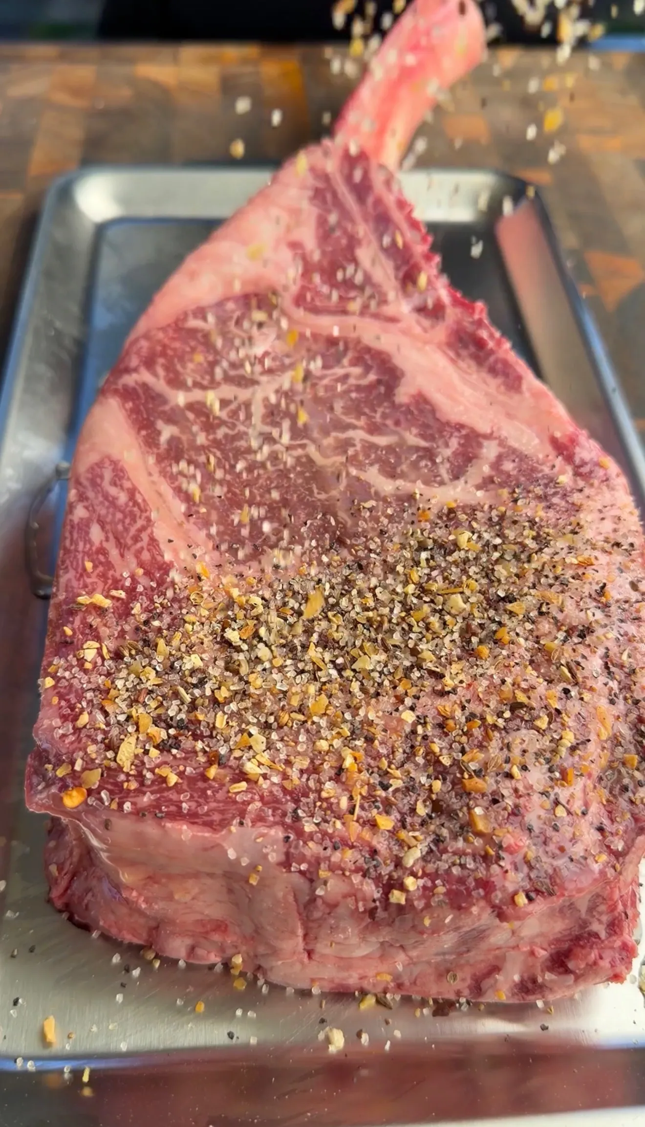 Raw tomahawk steak seasoned with spices and black pepper on a metal baking tray ready for grilling