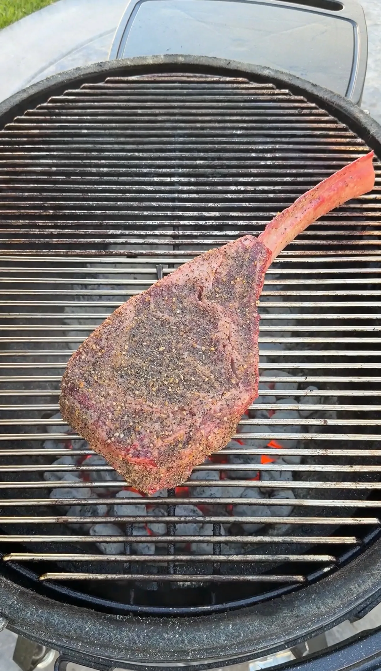 Large seasoned tomahawk steak with long bone handle cooking on round charcoal grill grates over glowing coals
