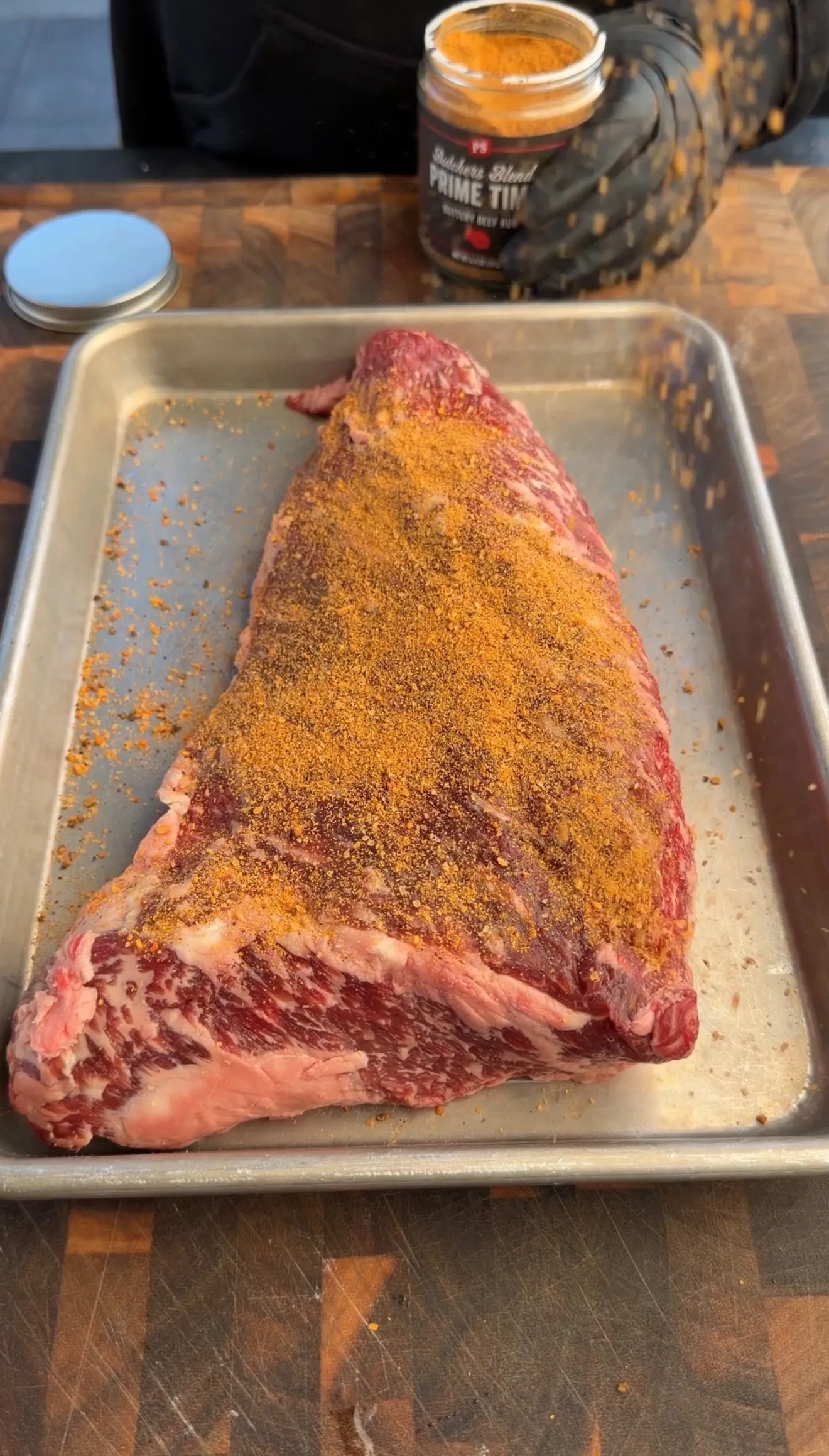 Raw tri-tip roast coated with orange spice rub on aluminum baking sheet with seasoning jar in background
