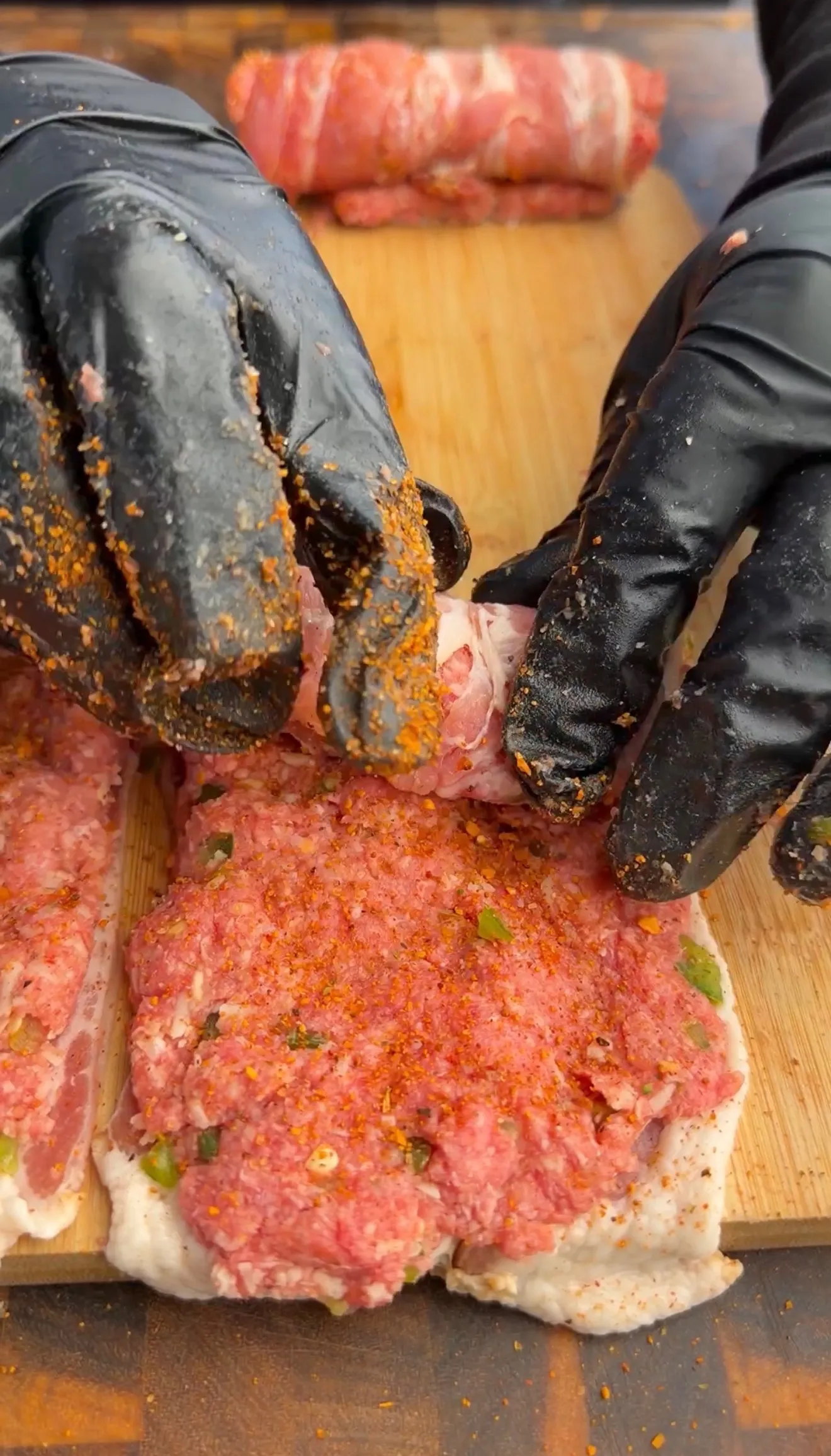 Hands in black gloves seasoning raw bacon-wrapped jalapeño beef skewers on wooden cutting board during preparation
