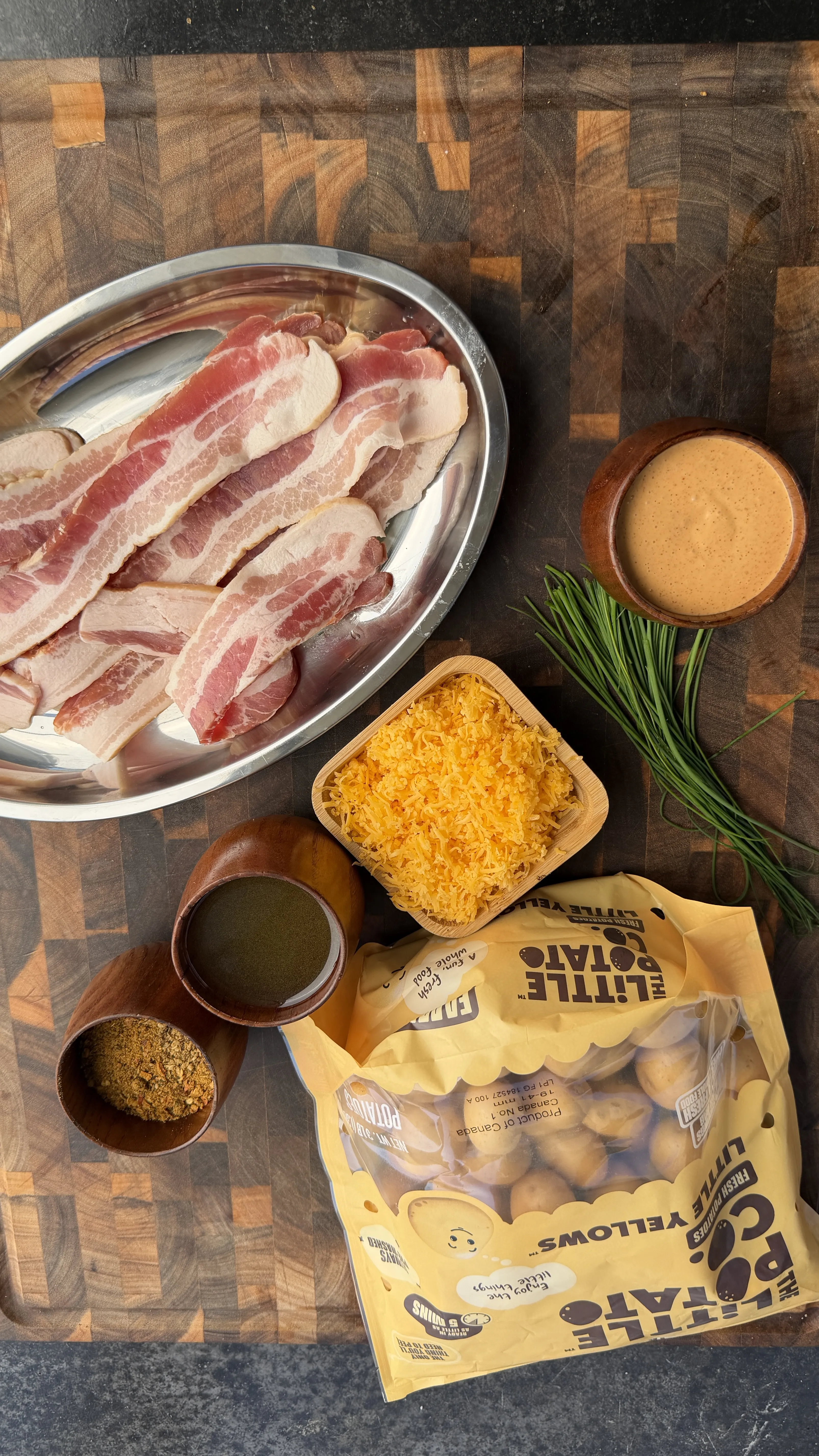 Raw bacon strips, grated cheese, fresh herbs, and baby potatoes arranged on wooden cutting board for loaded potato recipe