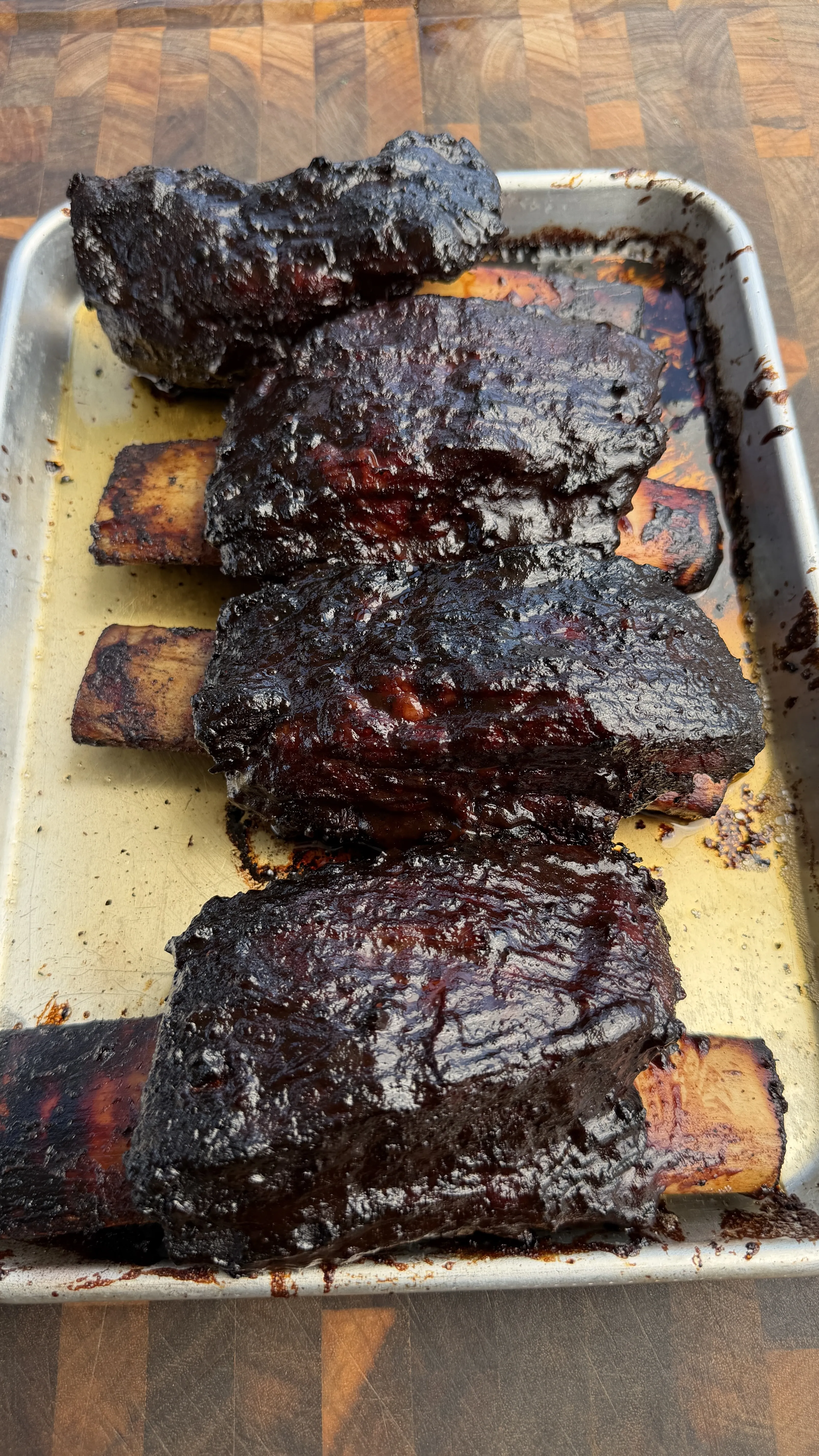 Dark smoky barbecue beef ribs with charred exterior in aluminum baking pan on wooden surface