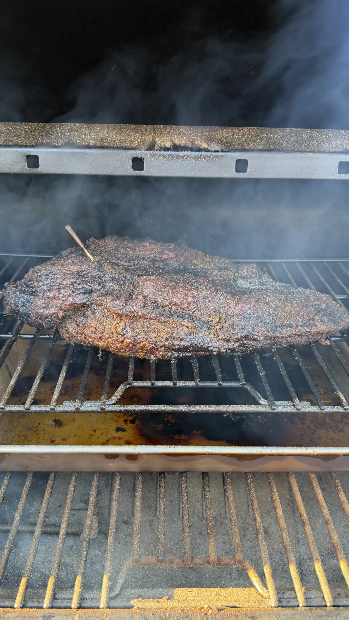 Large smoked beef brisket with dark bark crust cooking on barbecue grill grates with visible smoke
