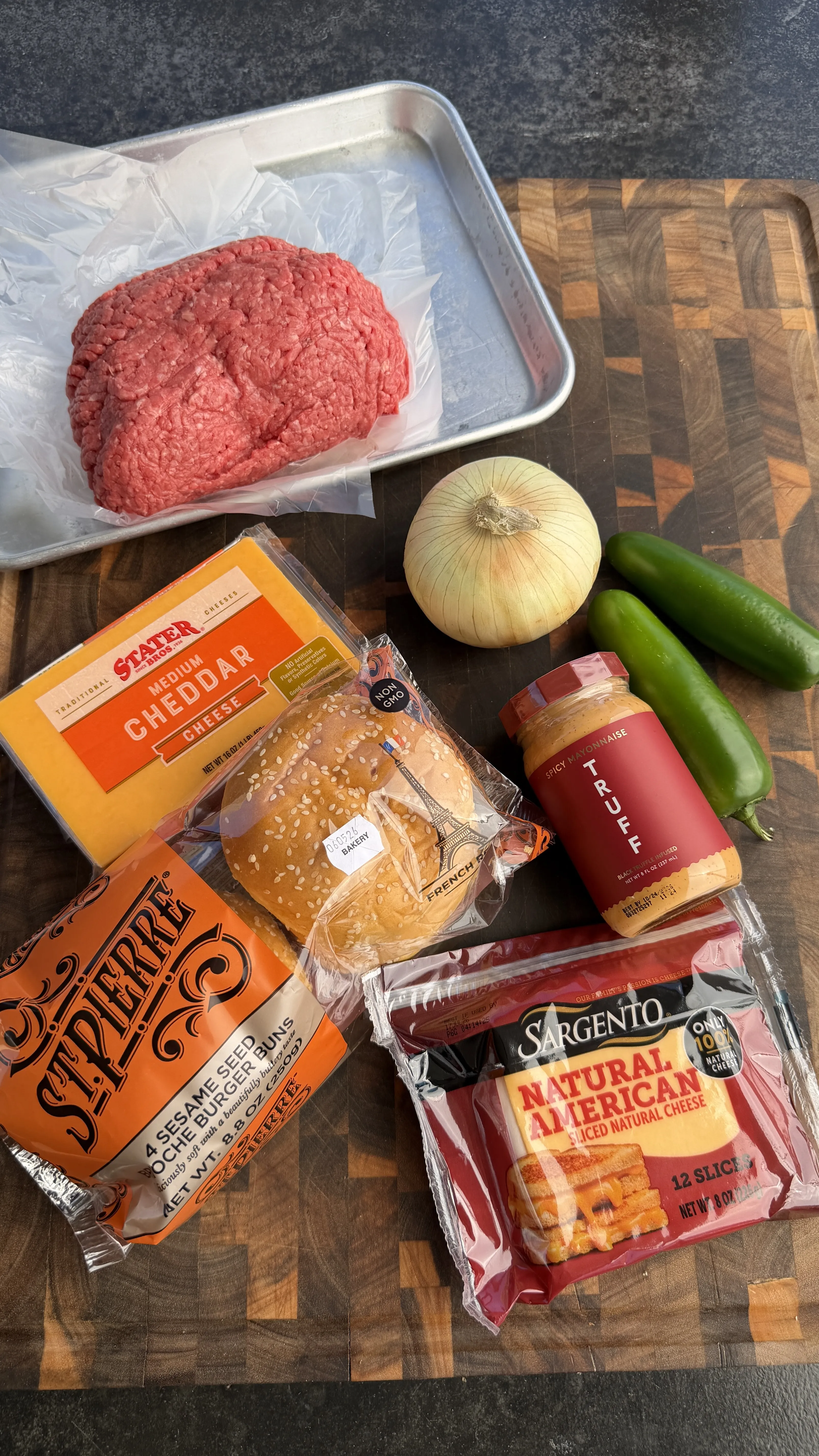 Raw ground beef, cheddar cheese, sesame seed buns, jalapeños, and onion arranged on wooden cutting board
