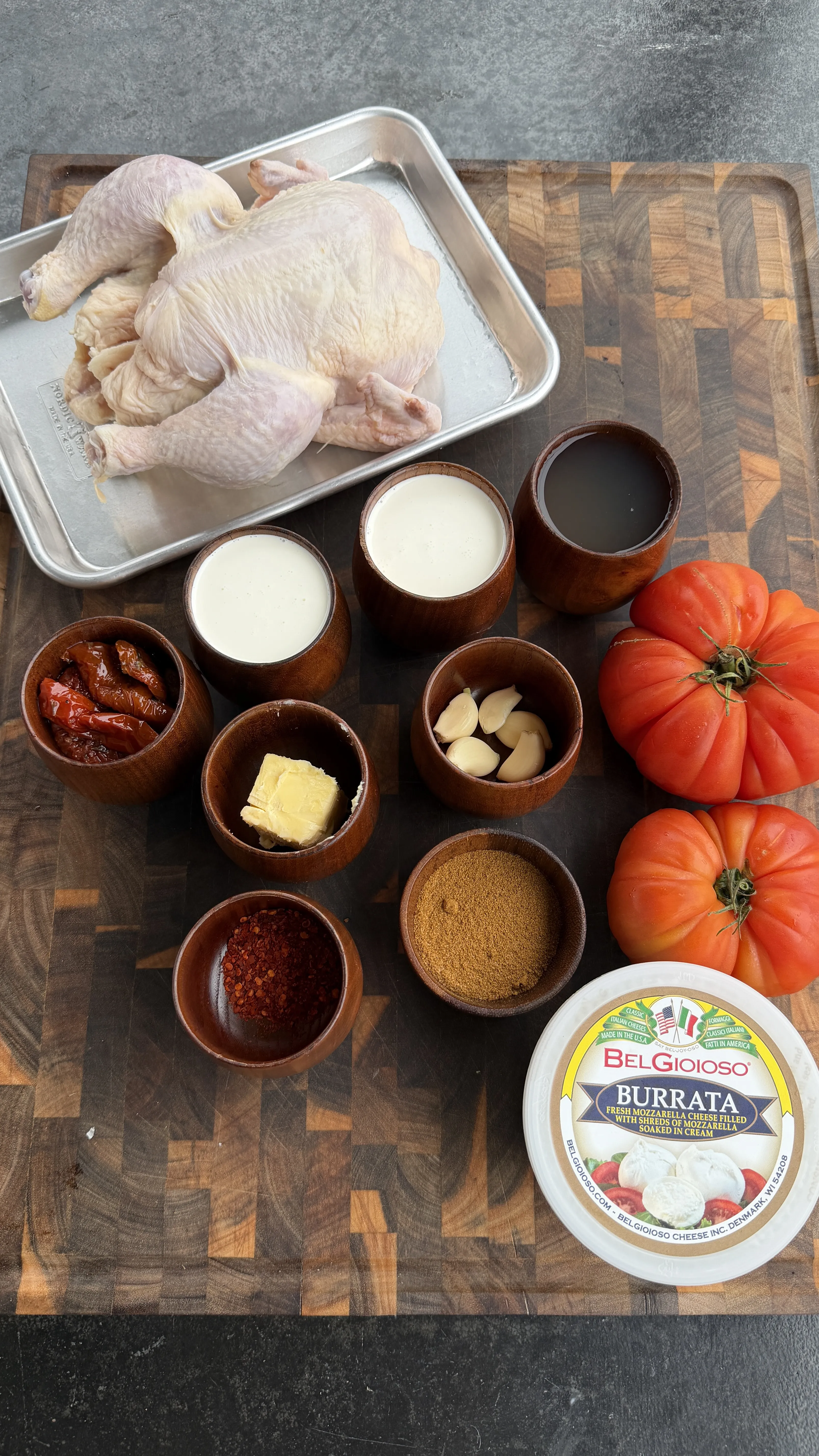 Raw spatchcock chicken with cooking ingredients including burrata cheese, fresh tomatoes, and spices in wooden bowls