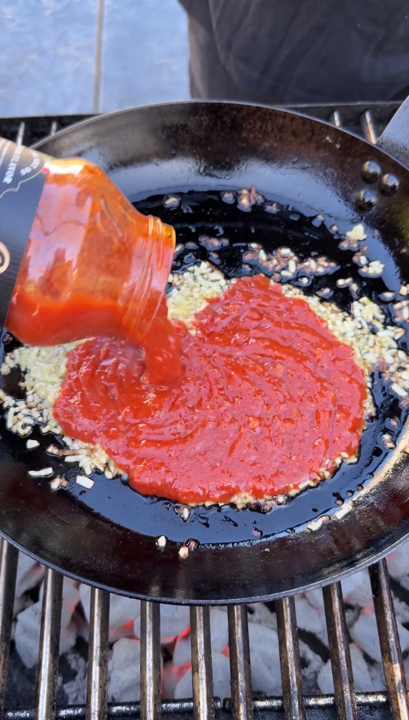 Red tomato sauce being poured from bottle into cast iron skillet with saut&eacute;ed garlic and onions