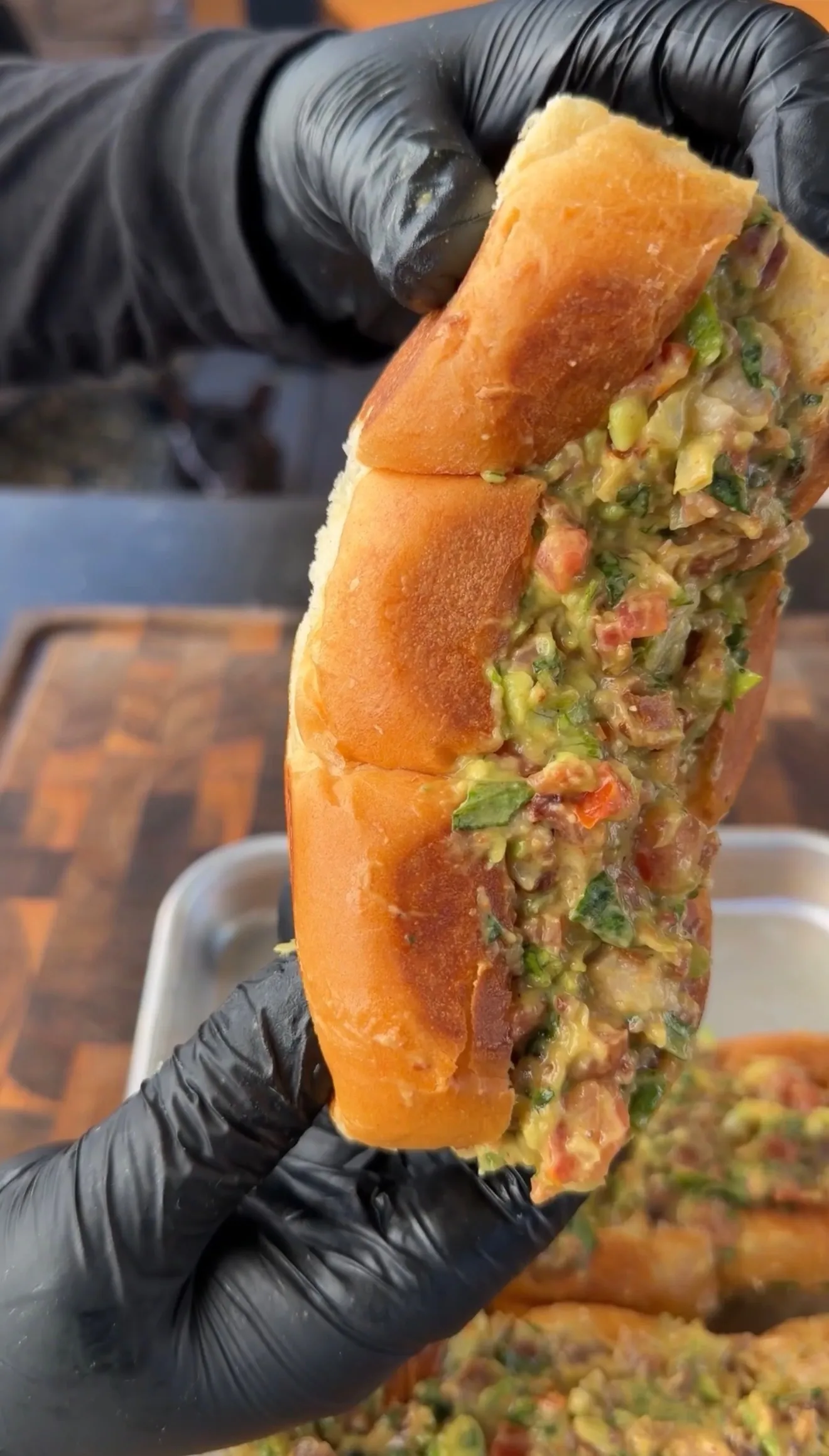 Hands in black gloves holding open a crusty slider roll stuffed with chunky avocado BLT filling