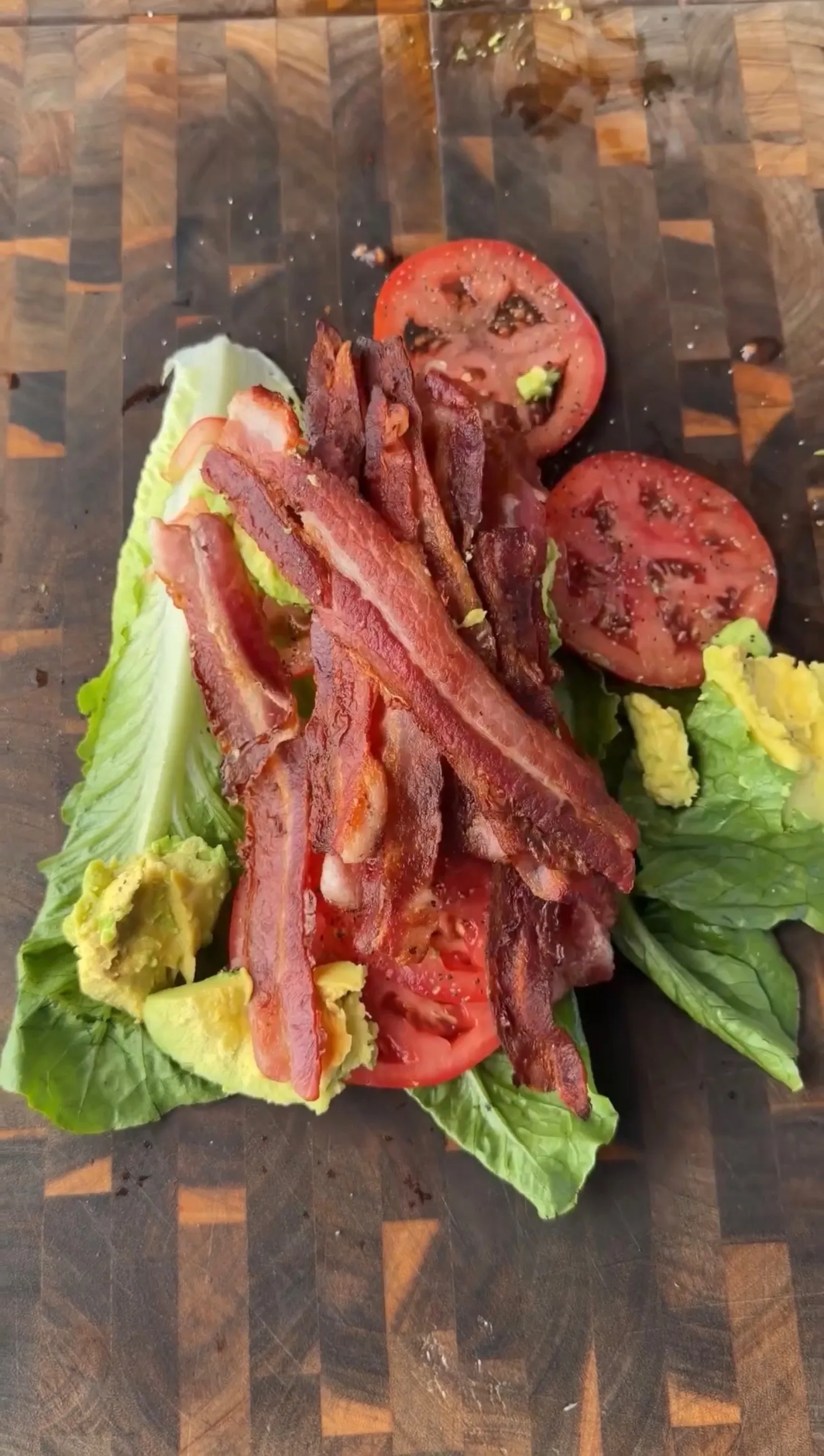 Avocado BLT lettuce wrap with crispy bacon strips, sliced tomatoes, and mashed avocado on a wooden cutting board