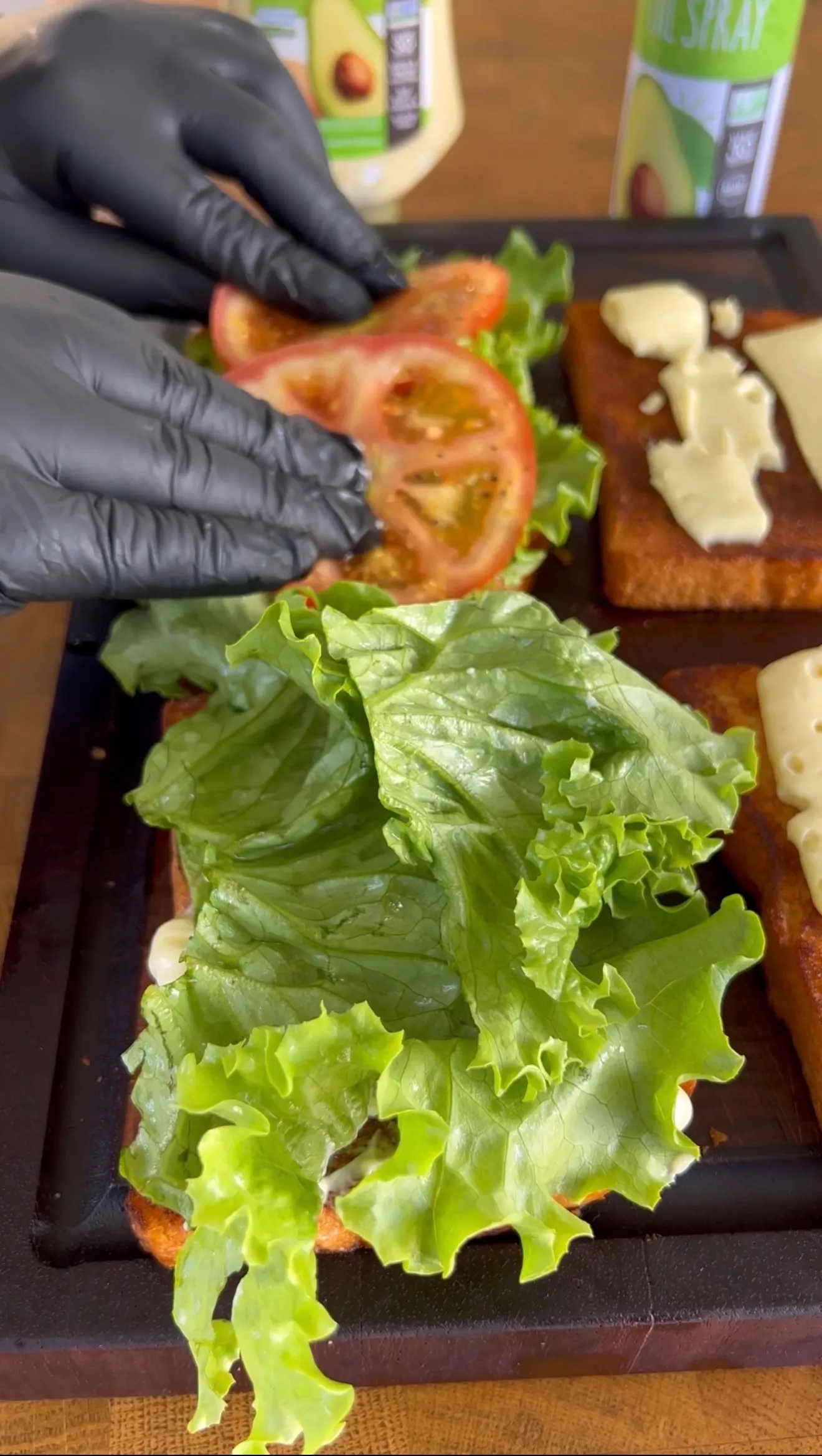 Hands in black gloves assembling an Avocado BLT sandwich with fresh lettuce, tomato slices, and ingredients on board