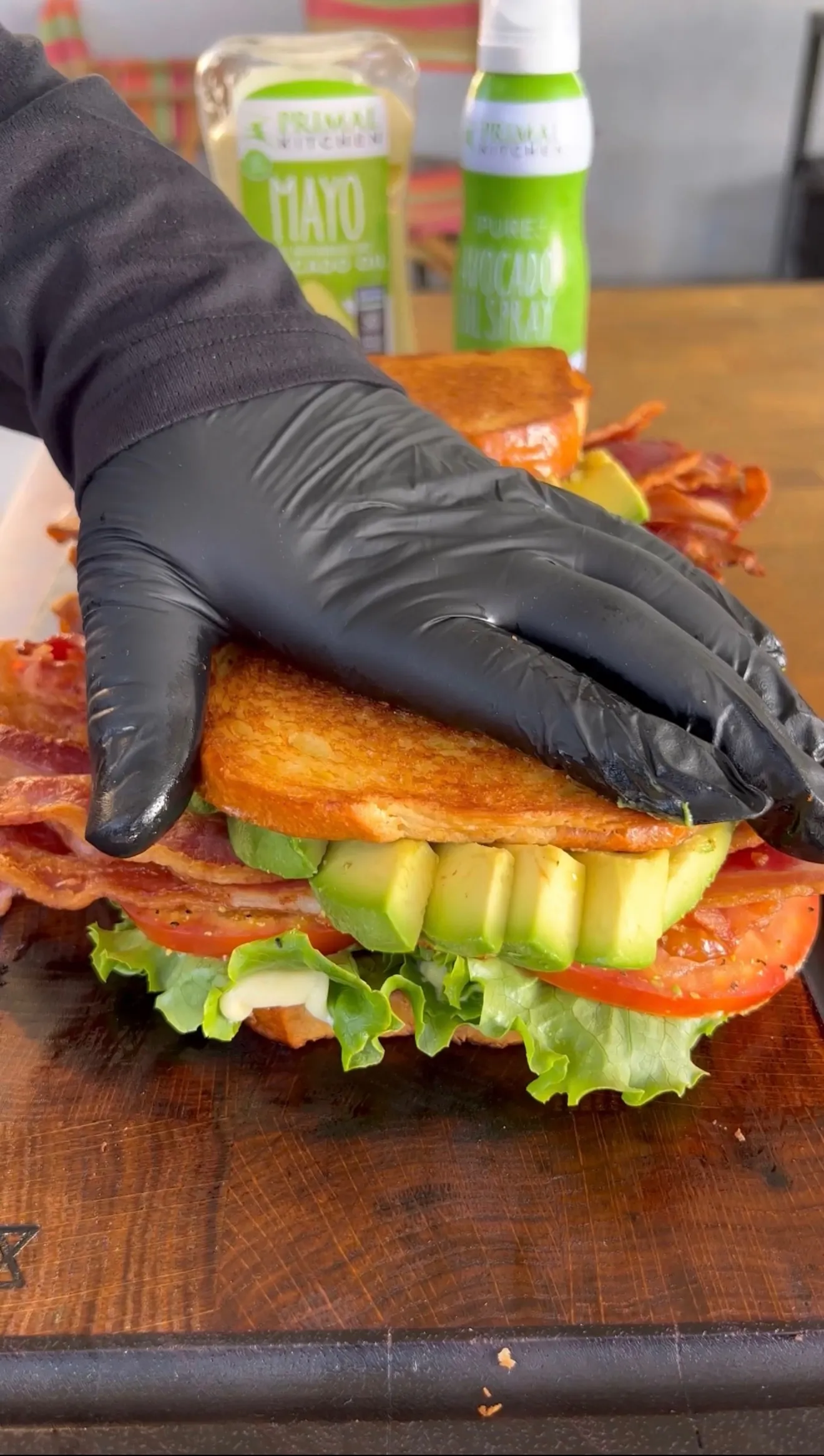 Hands in black gloves assembling an avocado BLT sandwich with bacon, lettuce, tomato on wooden cutting board