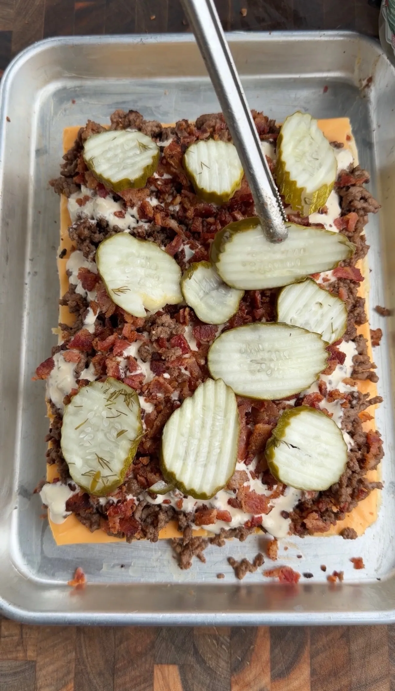 Bacon cheeseburger slider casserole with ground beef, bacon, cheese, and pickle slices being assembled in aluminum pan