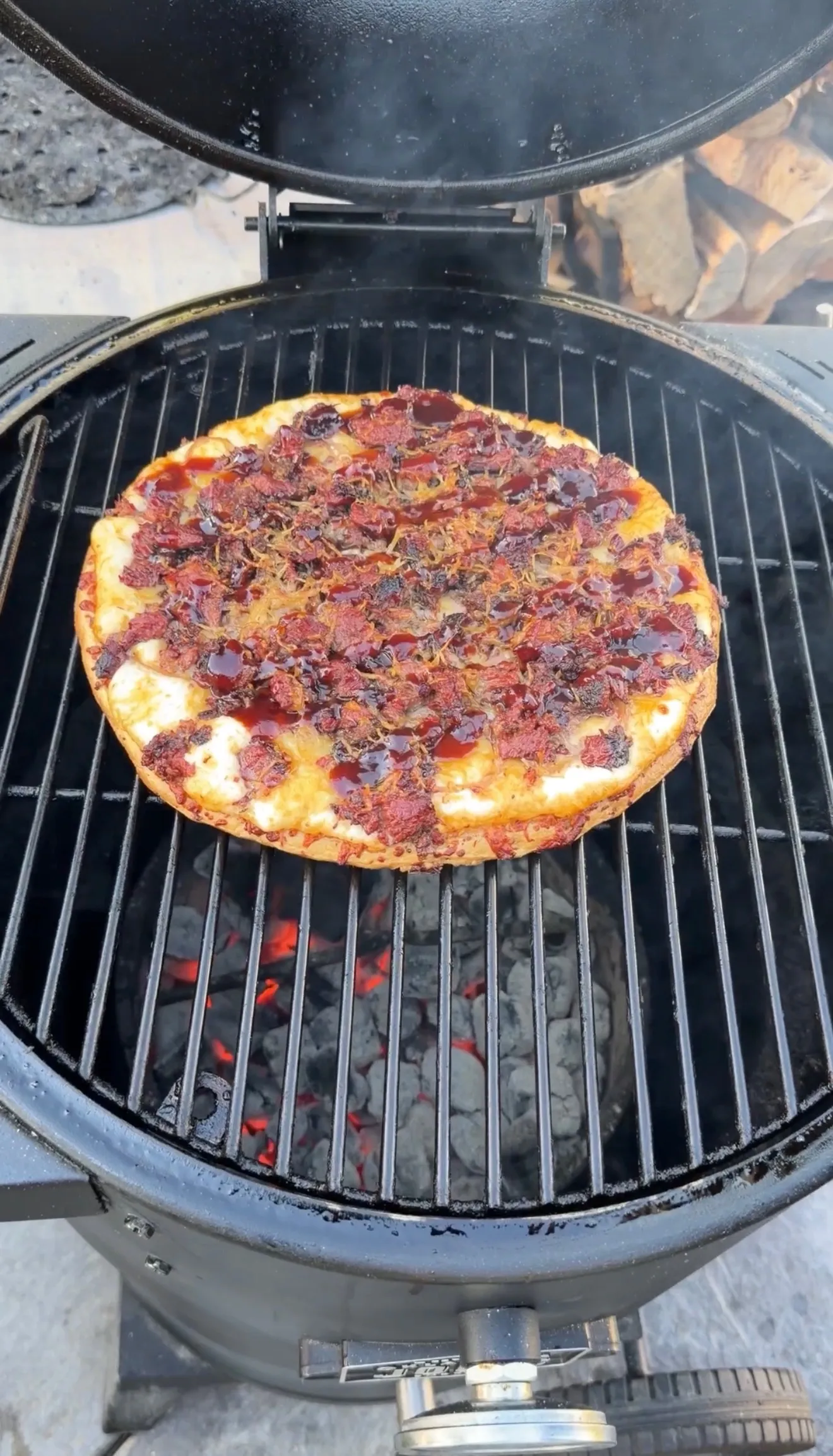 Barbecue brisket pizza with melted cheese cooking on a round kettle grill with glowing coals beneath