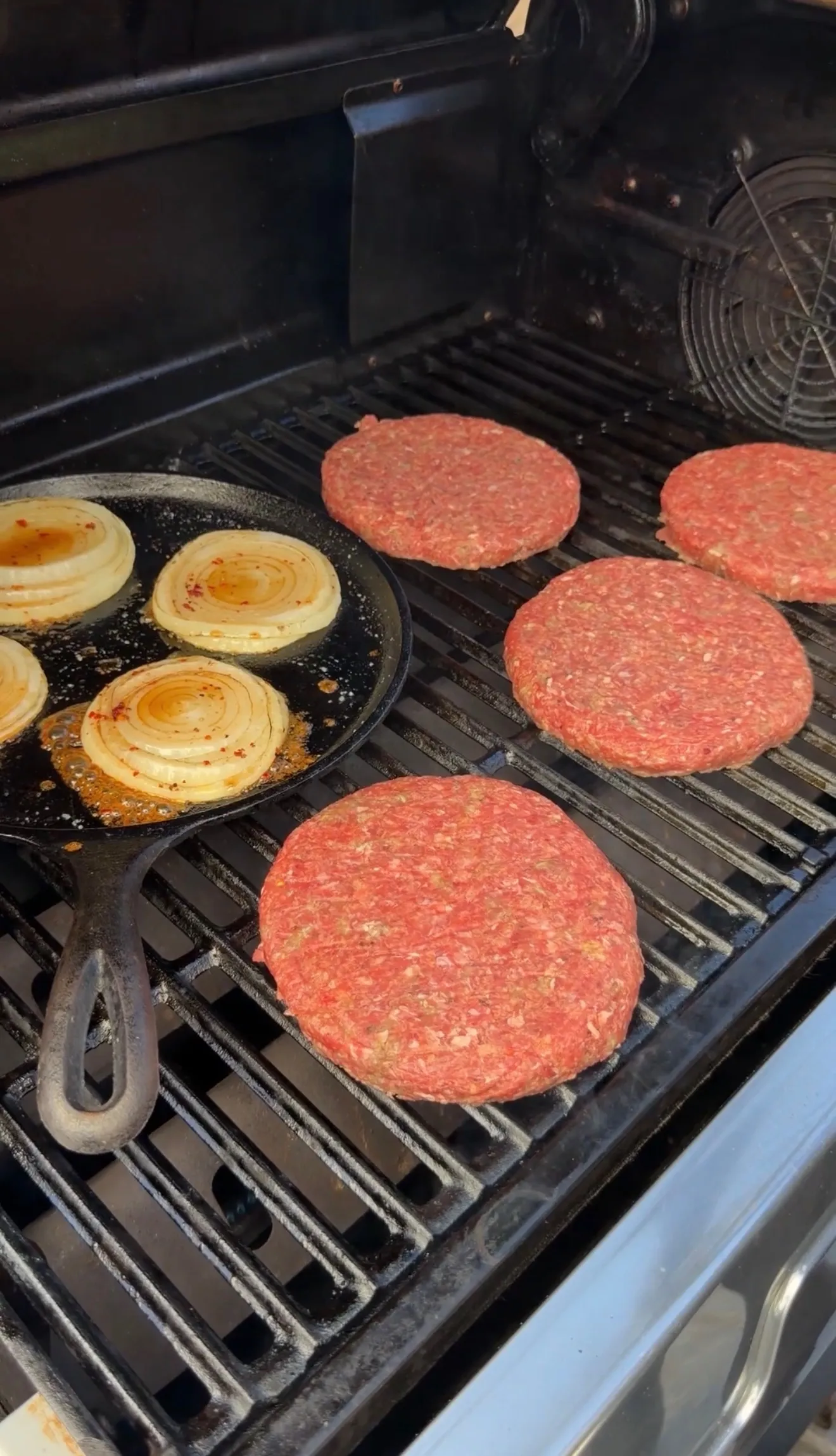 Raw bison burger patties cooking on grill grates alongside caramelized onions in cast iron skillet