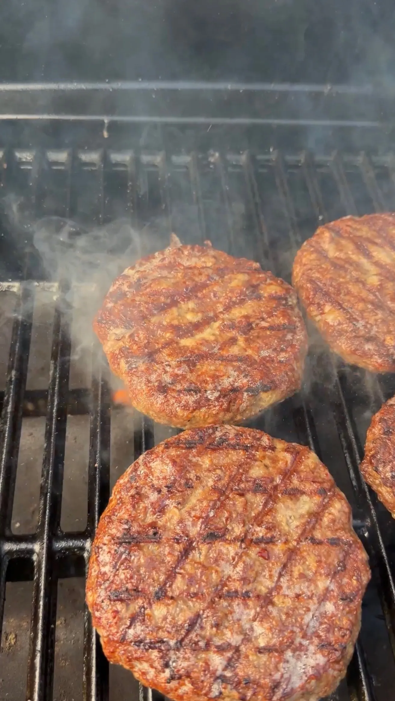 Four bourbon glazed bison burger patties cooking on a smoking barbecue grill with distinctive char marks