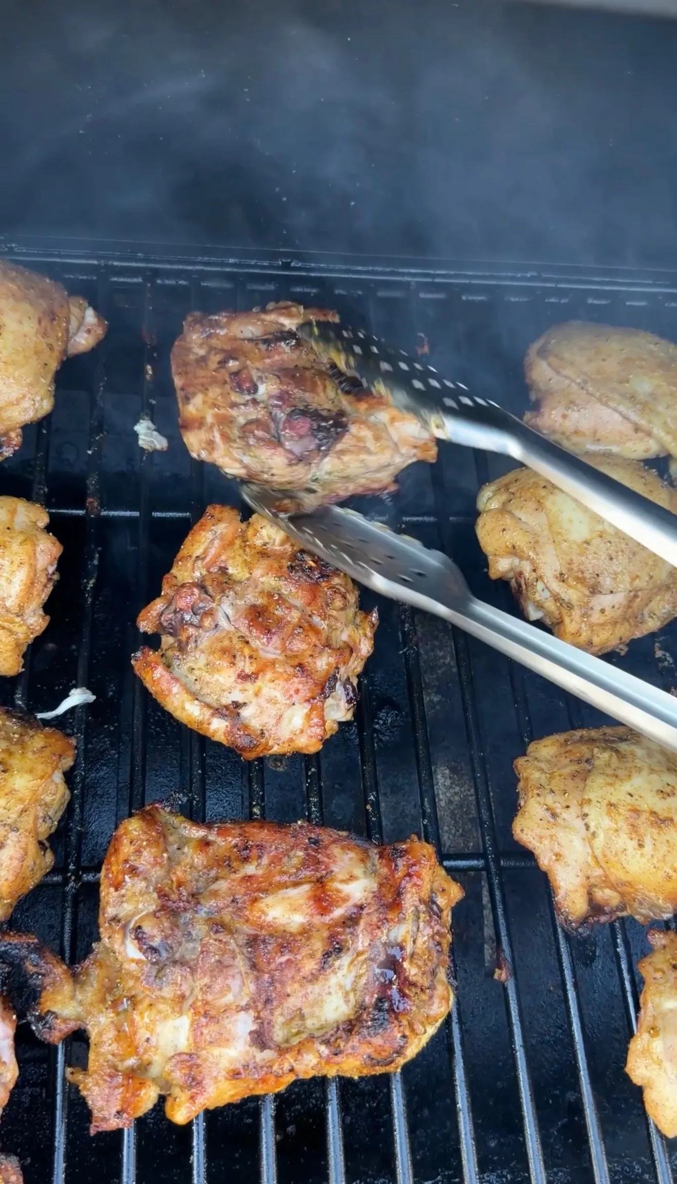Cajun lemon pepper honey chicken thighs grilling on a smoky BBQ grill, being flipped with metal tongs