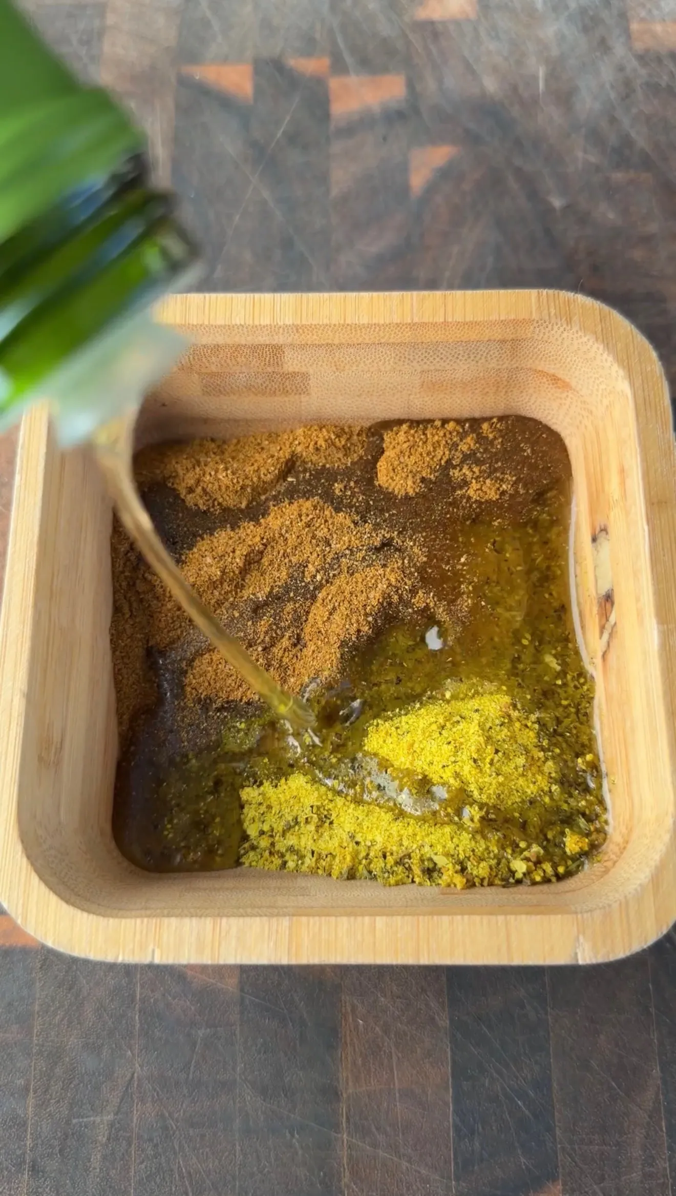 Olive oil being poured over Cajun and lemon pepper spices in a square wooden bowl for chicken marinade