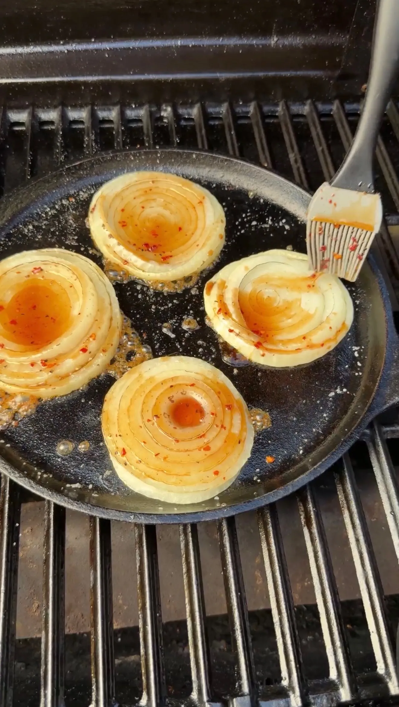Four caramelized onion halves cooking in a cast iron skillet on a grill with basting brush