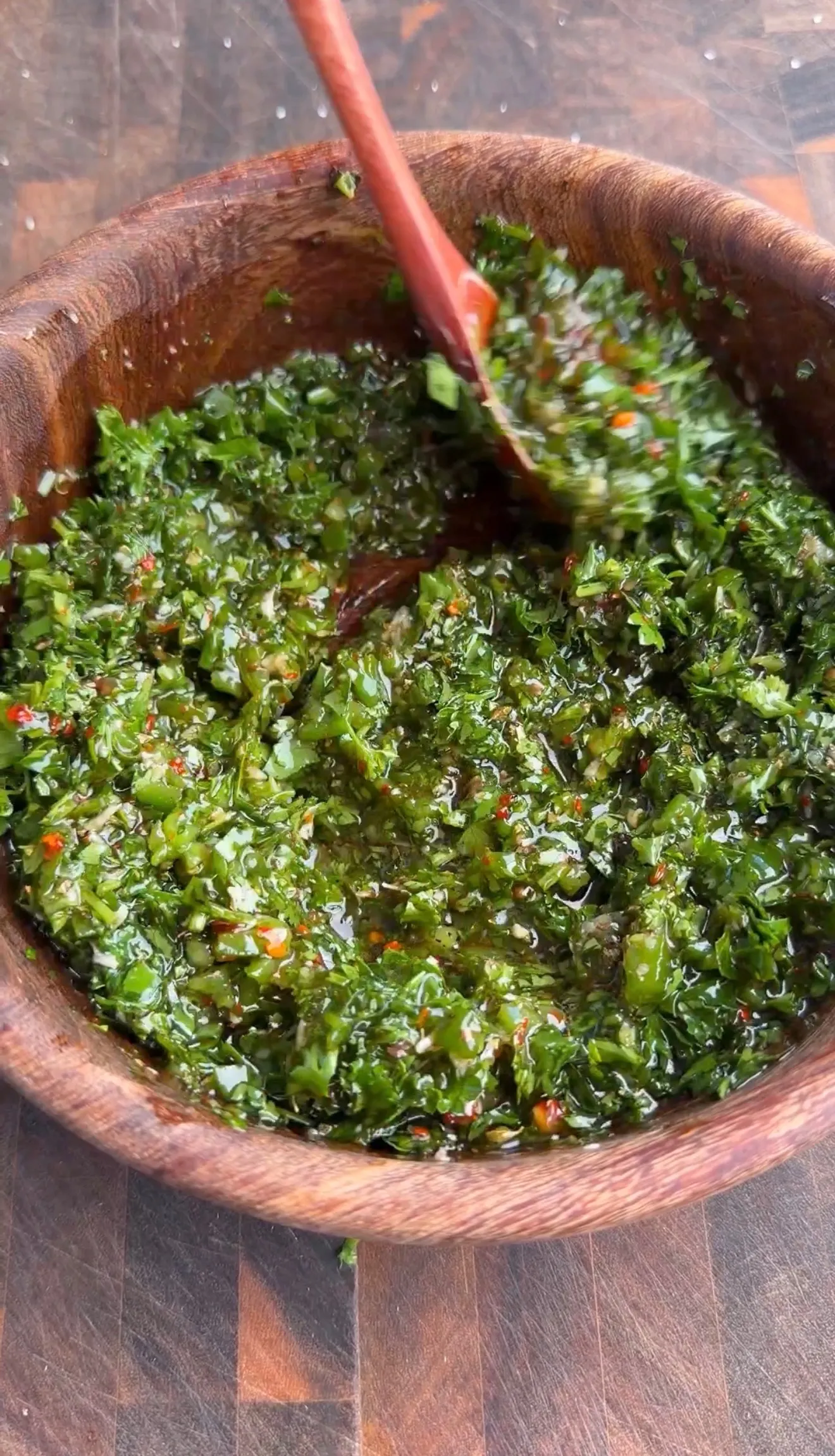 Fresh charred jalapeño chimichurri with herbs and spices being mixed in a wooden bowl with red spoon