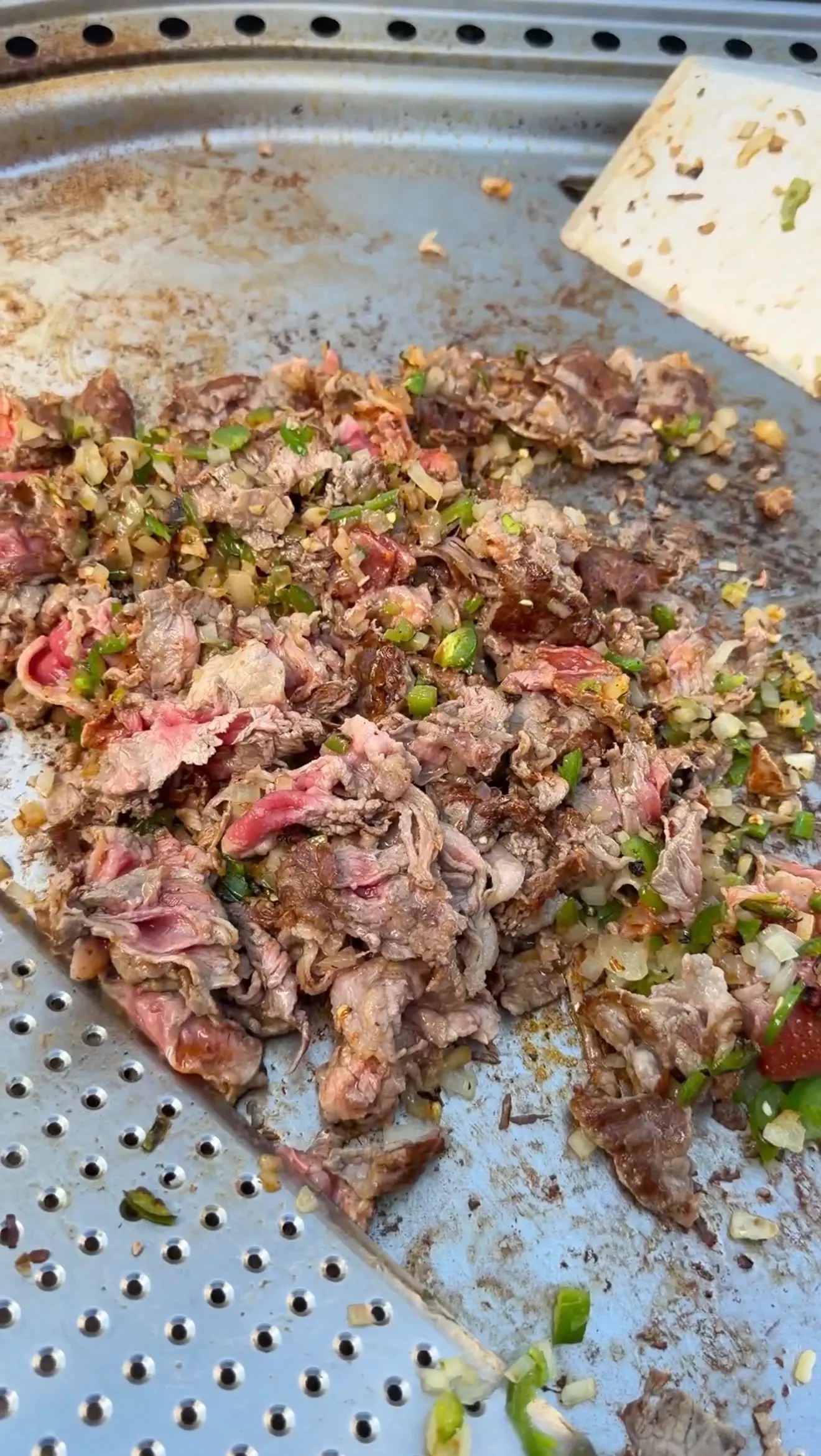 Chopped cheesesteak meat with green onions and garlic cooking on a perforated metal griddle pan