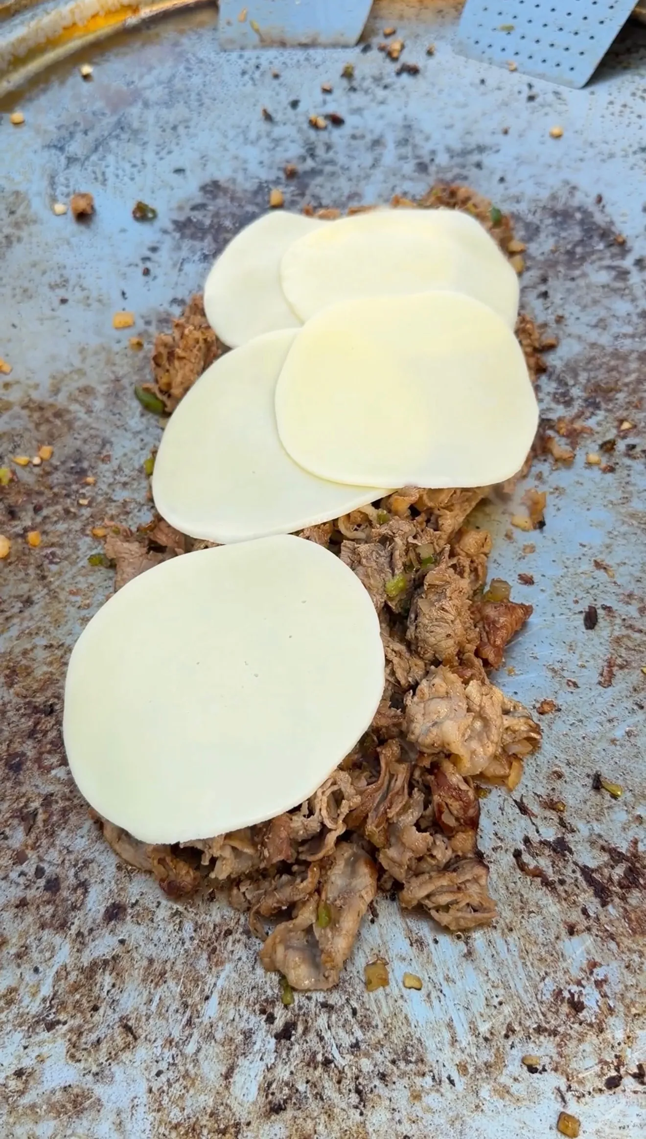 Seasoned cheesesteak meat with peppers topped with melting provolone cheese slices on a flat iron griddle