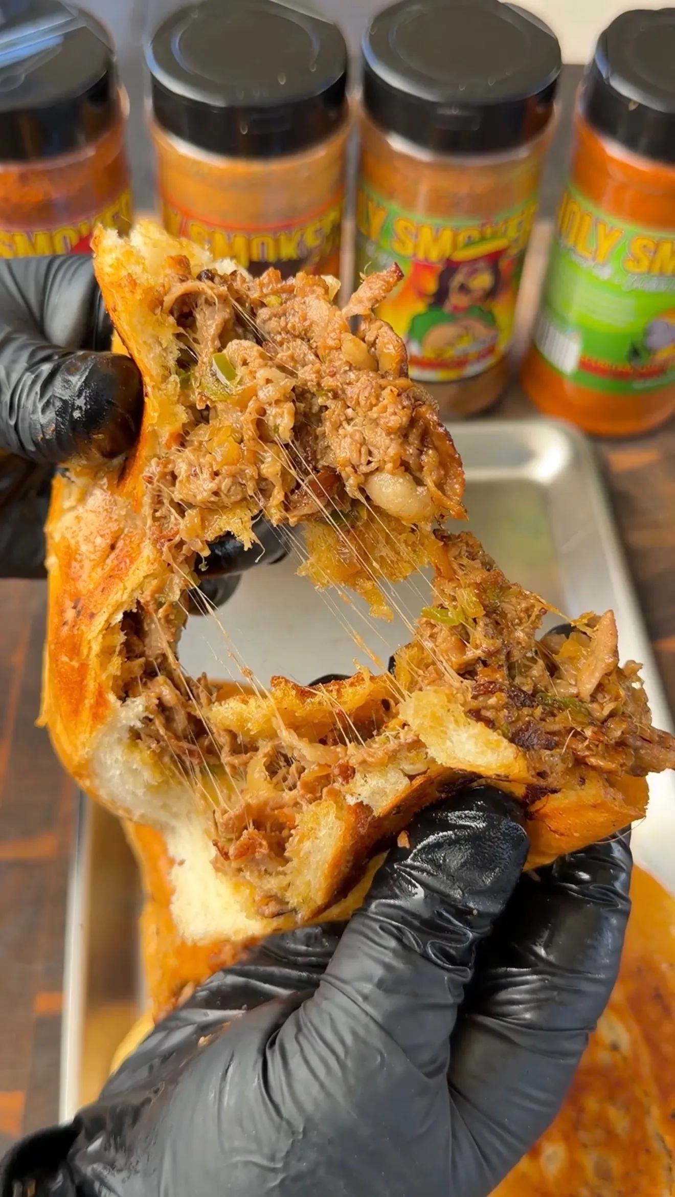 Hands pulling apart cheesy cheesesteak stuffed bolillo bread showing melted cheese pull with seasoning jars in background