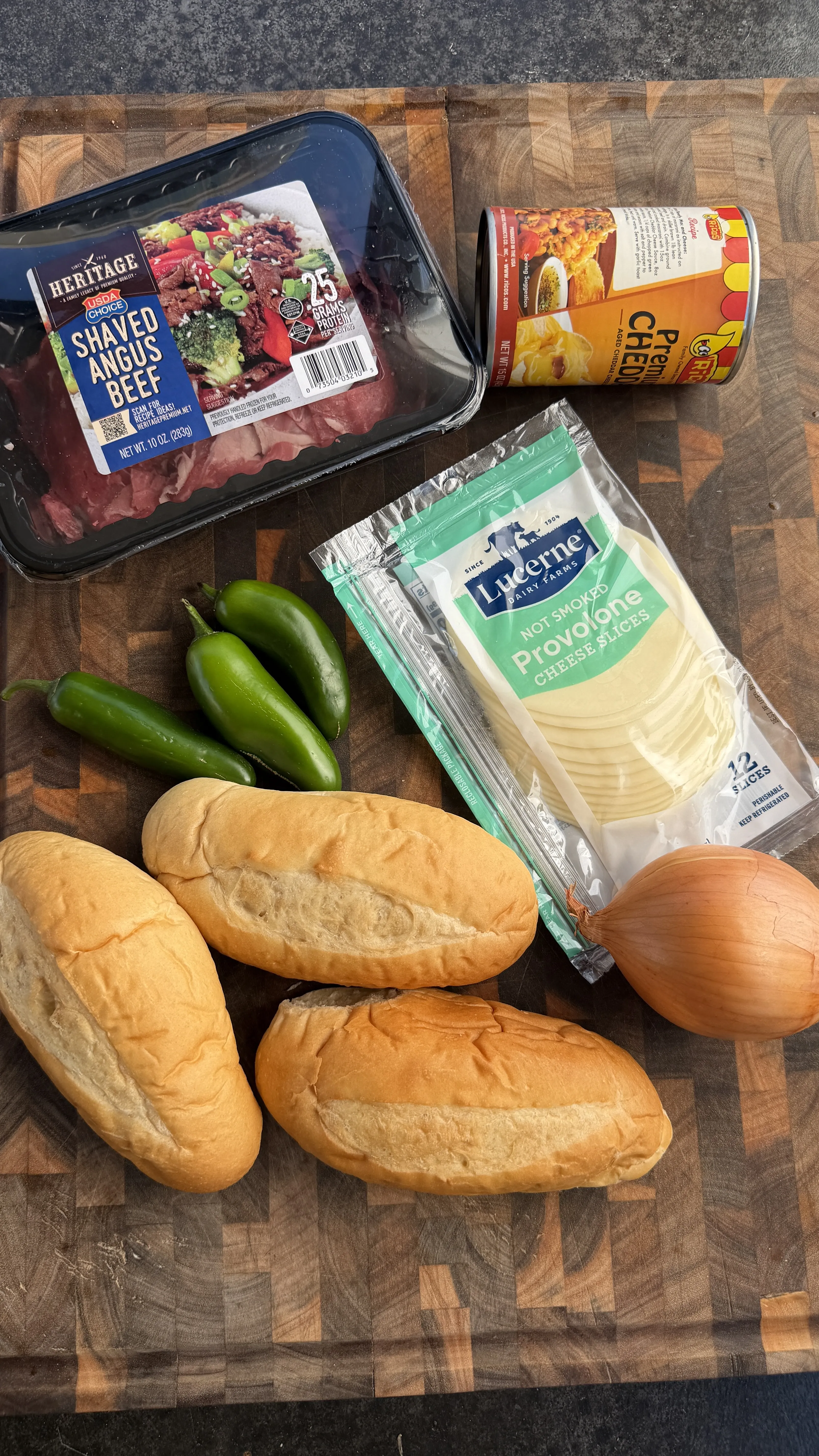 Cheesesteak stuffed bolillo ingredients: shaved angus beef, provolone, jalapeños, onion, canned cheese, and rolls on cutting board