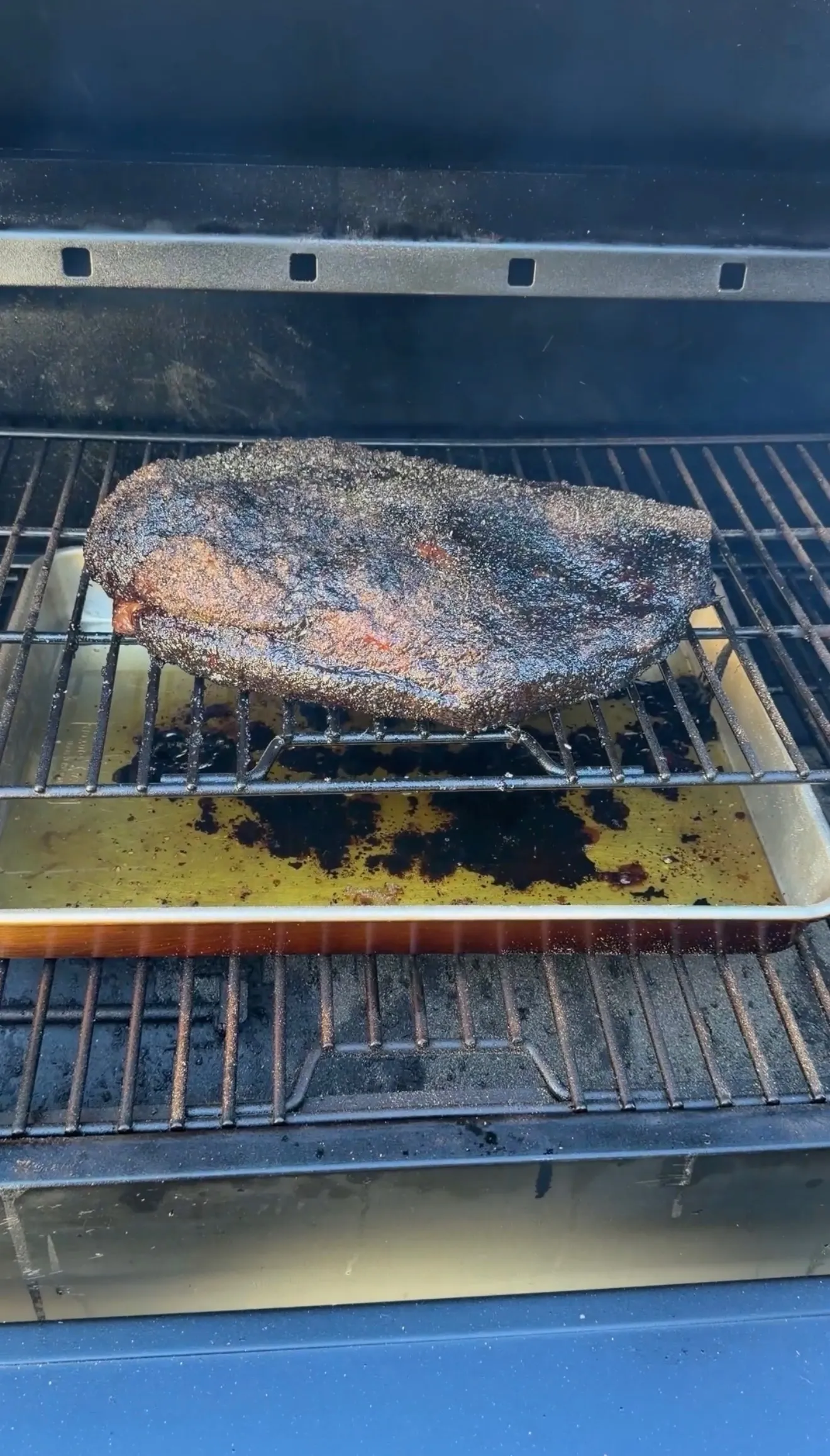 Whole beef brisket with dark bark crust smoking on a pellet grill rack with drip pan below