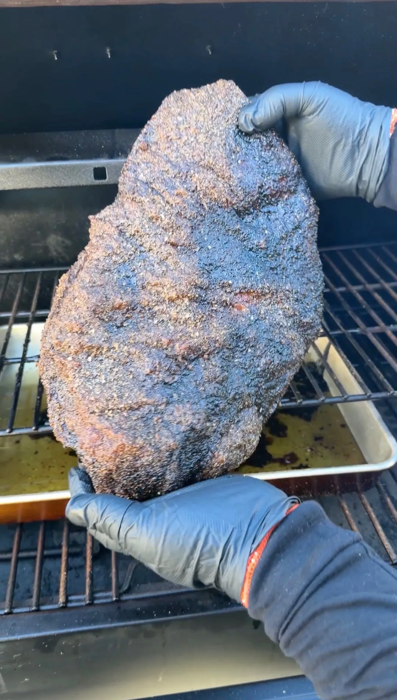 Hands in BBQ gloves lifting a whole smoked brisket with dark bark crust from a pellet smoker grill