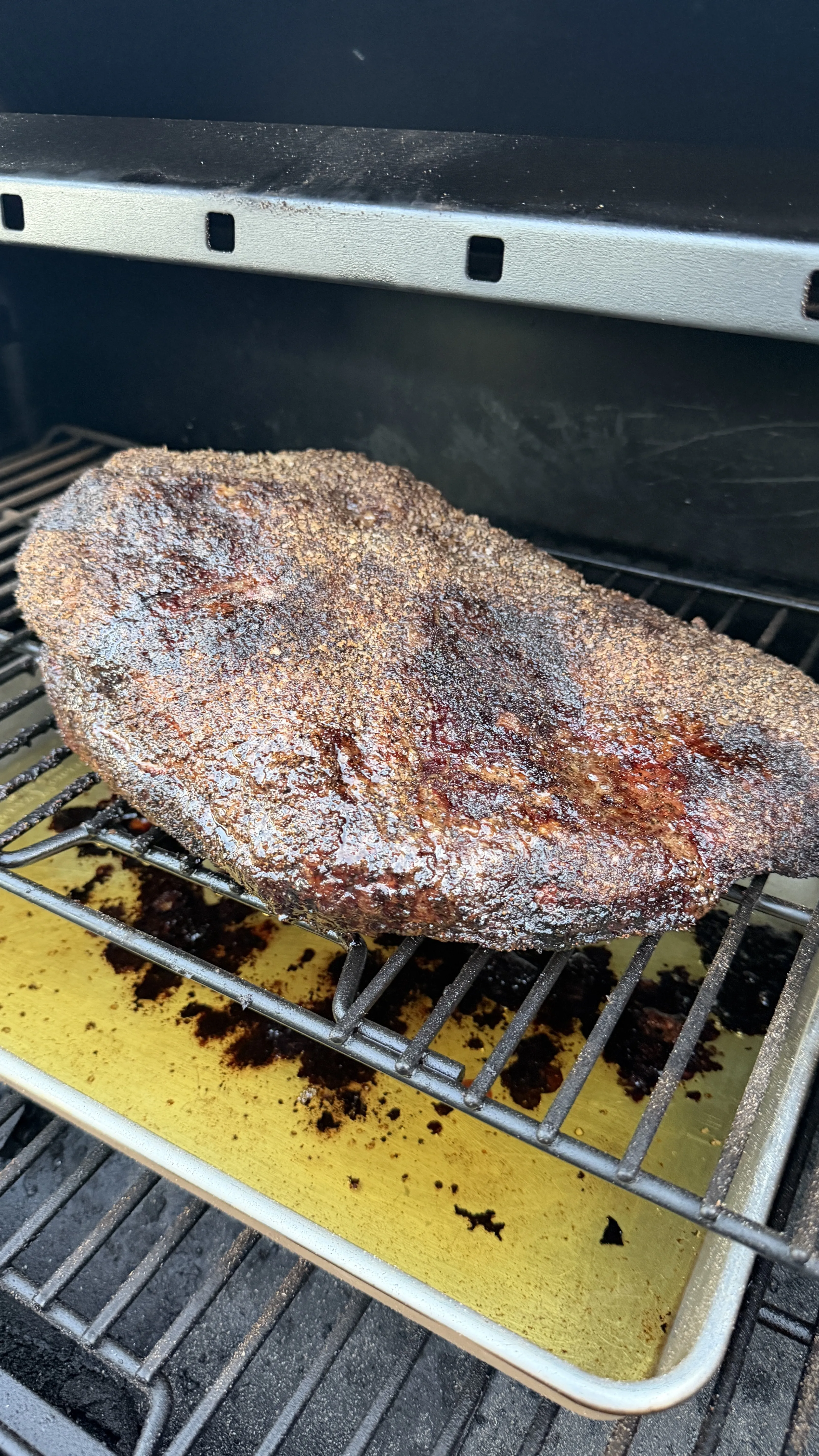 Whole beef brisket coated in dry rub smoking on a grill rack with a drip pan underneath