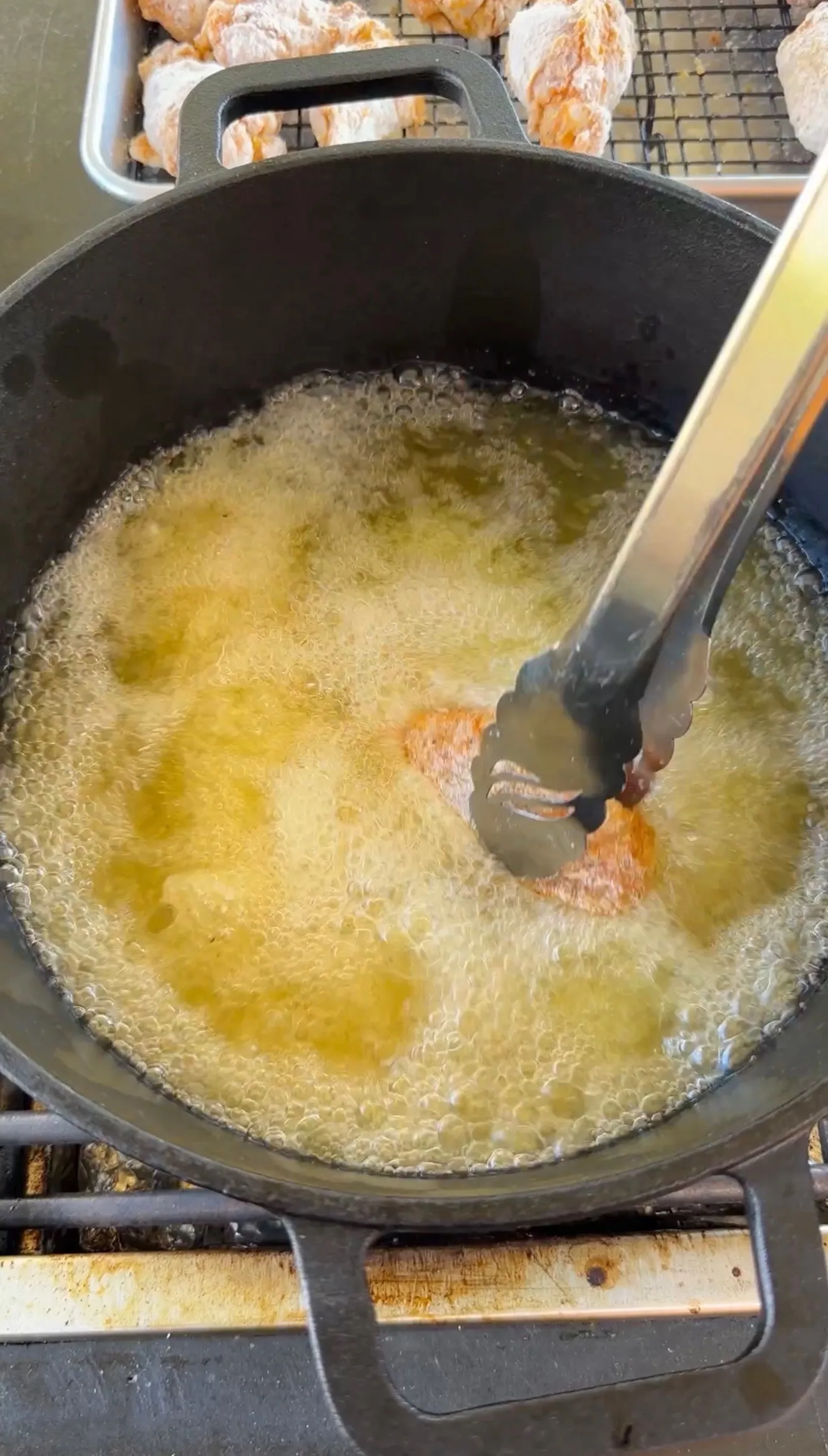 Chicken wings deep frying in bubbling hot oil in a cast iron Dutch oven with tongs, cooked wings resting on rack behind