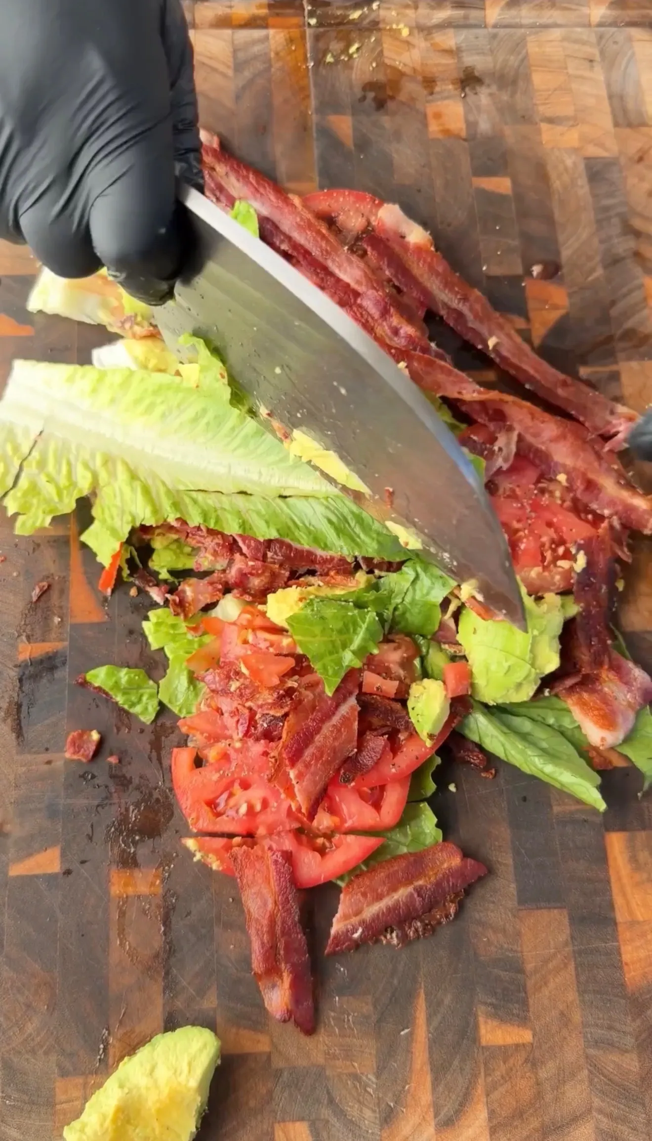 Gloved hand chopping bacon, tomato, lettuce, and avocado for BLT sliders on a wooden cutting board