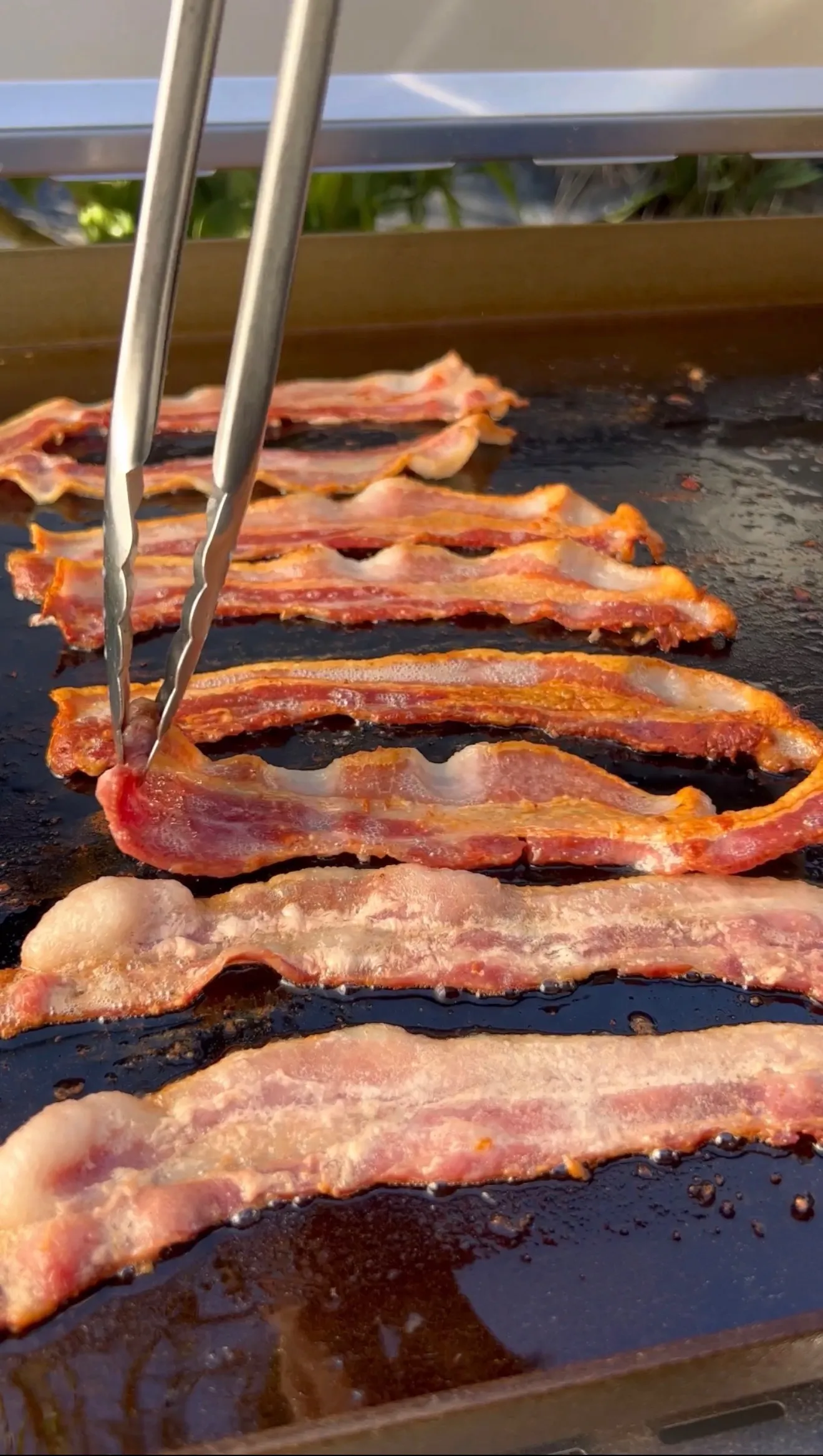 Crispy bacon strips cooking on a black griddle with tongs, showing various stages of doneness for BLT sandwich