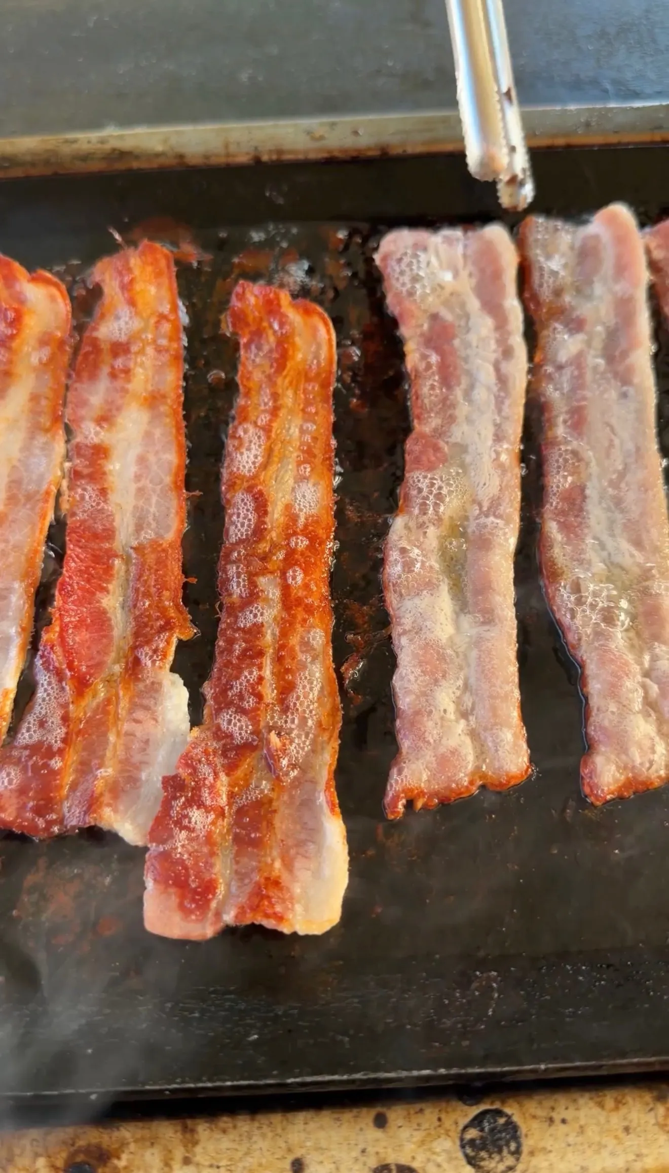 Strips of bacon sizzling and cooking on a flat iron griddle with tongs, some slices more cooked than others