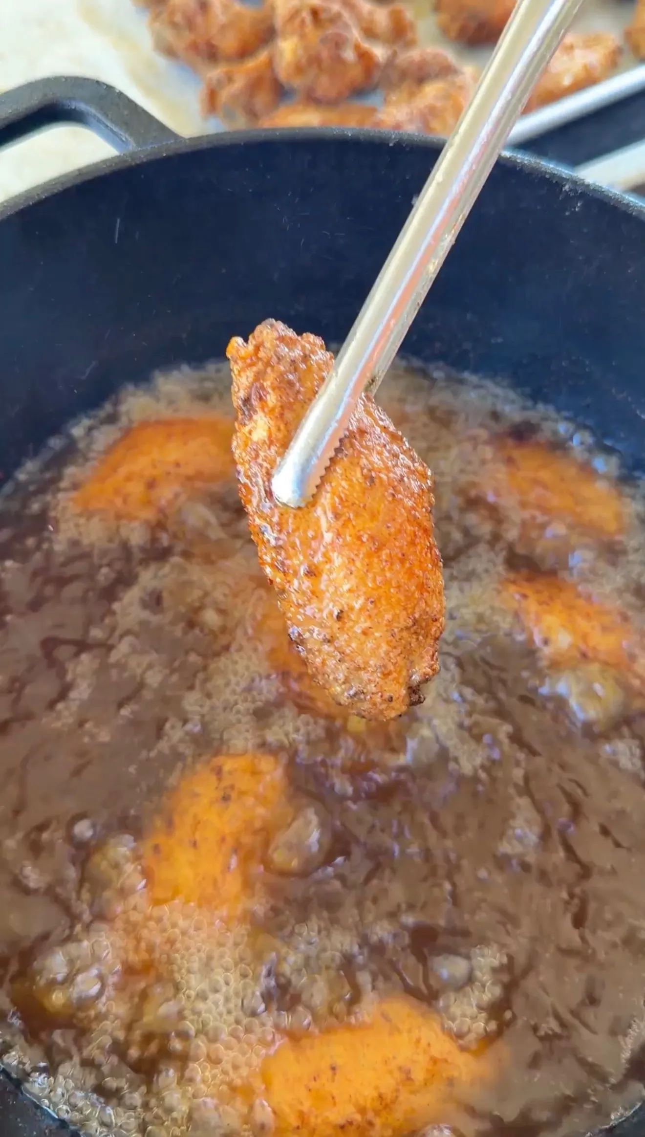 A crispy seasoned chicken wing held by tongs over bubbling hot oil in a cast iron skillet